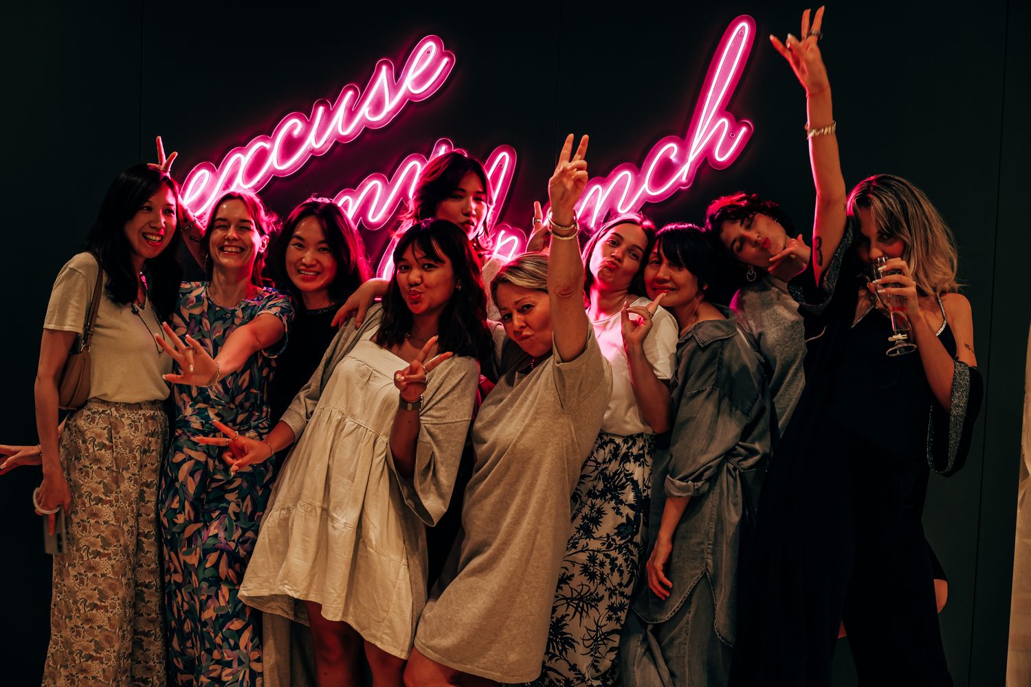 Group of women laughing and posing playfully beneath a vibrant pink neon sign.
