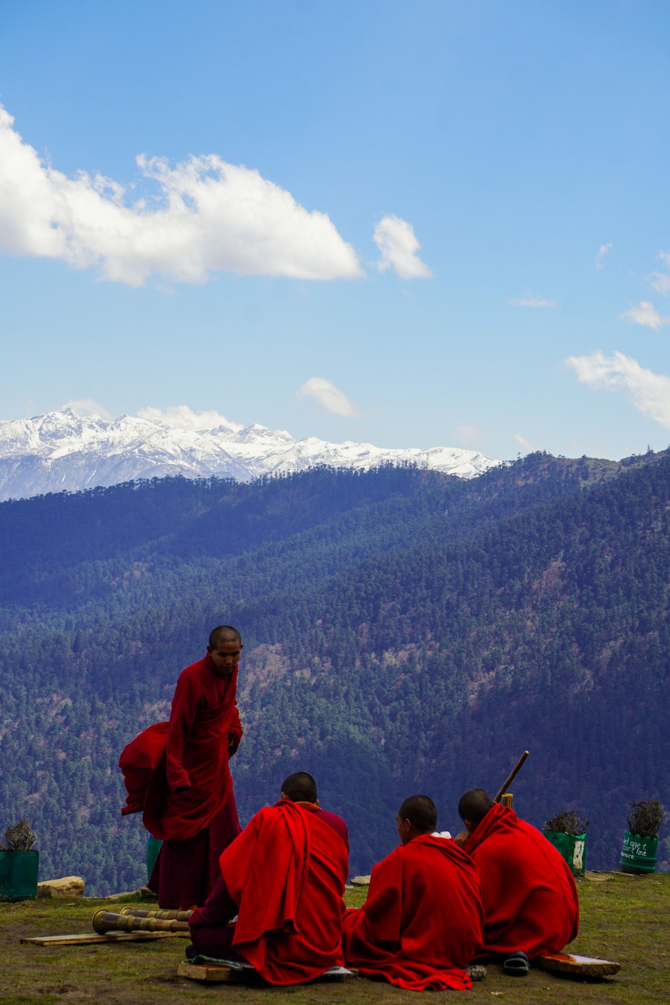 Buddhist monks in red robes gather on a hillside with stunning snow-capped Himalayan mountain views beyond.