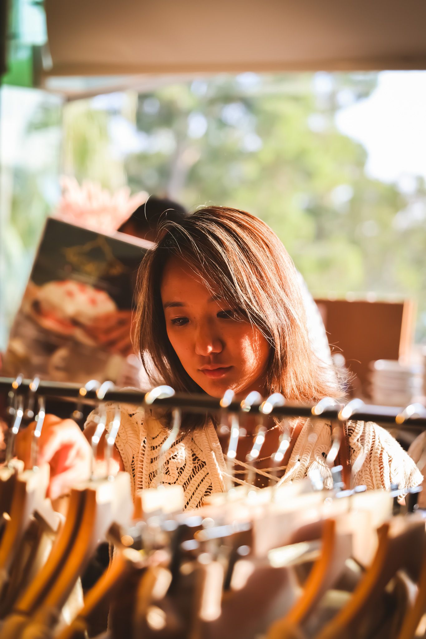 Young woman browsing through clothing rack in a sunlit boutique store with natural light background.