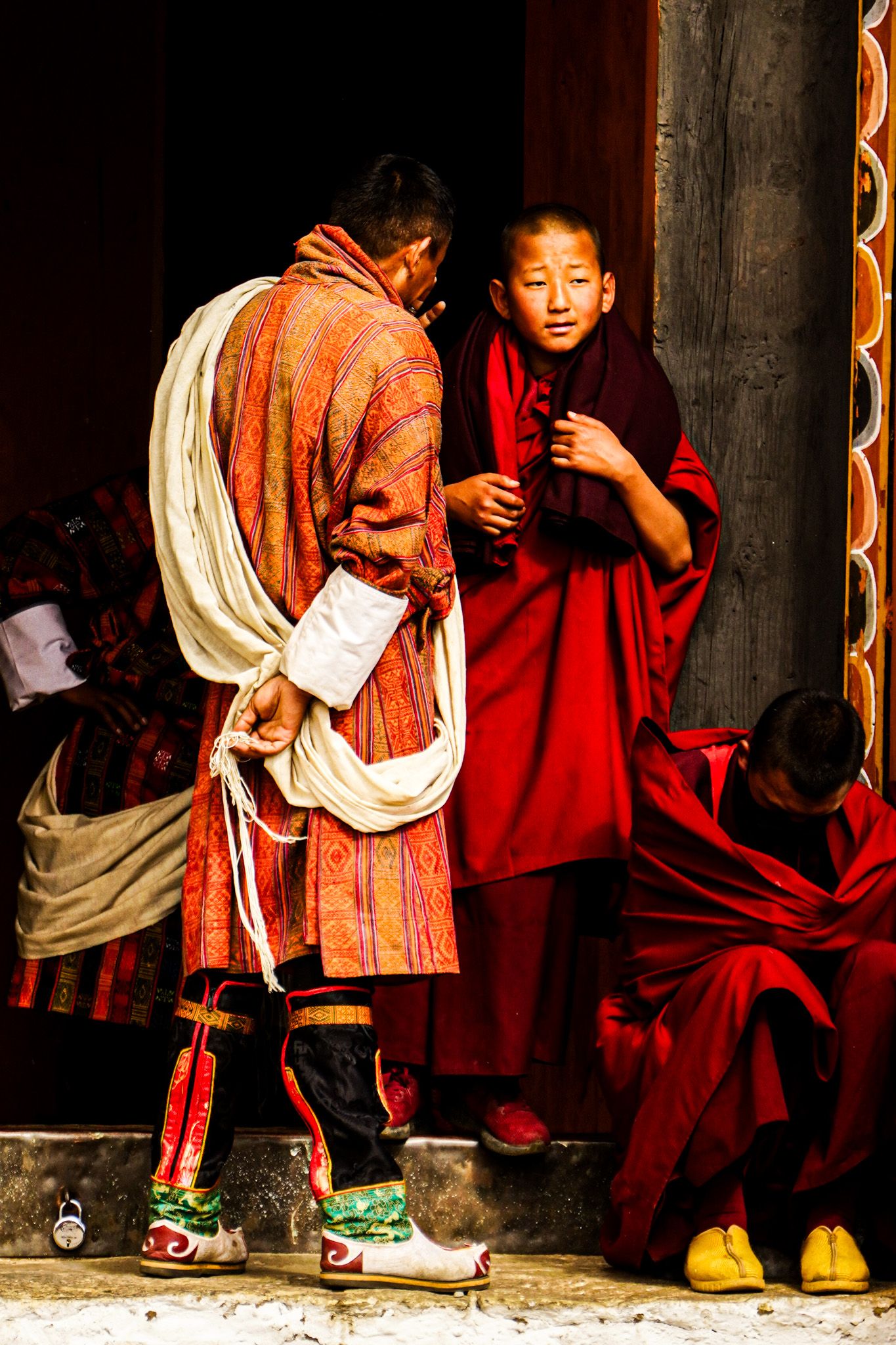 Young novice monk in red robes interacts with traditionally dressed Bhutanese man at temple steps.