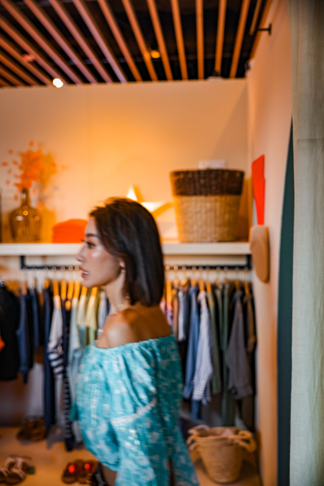 Woman in teal off-shoulder dress browsing clothing rack in a warmly lit boutique shop interior.