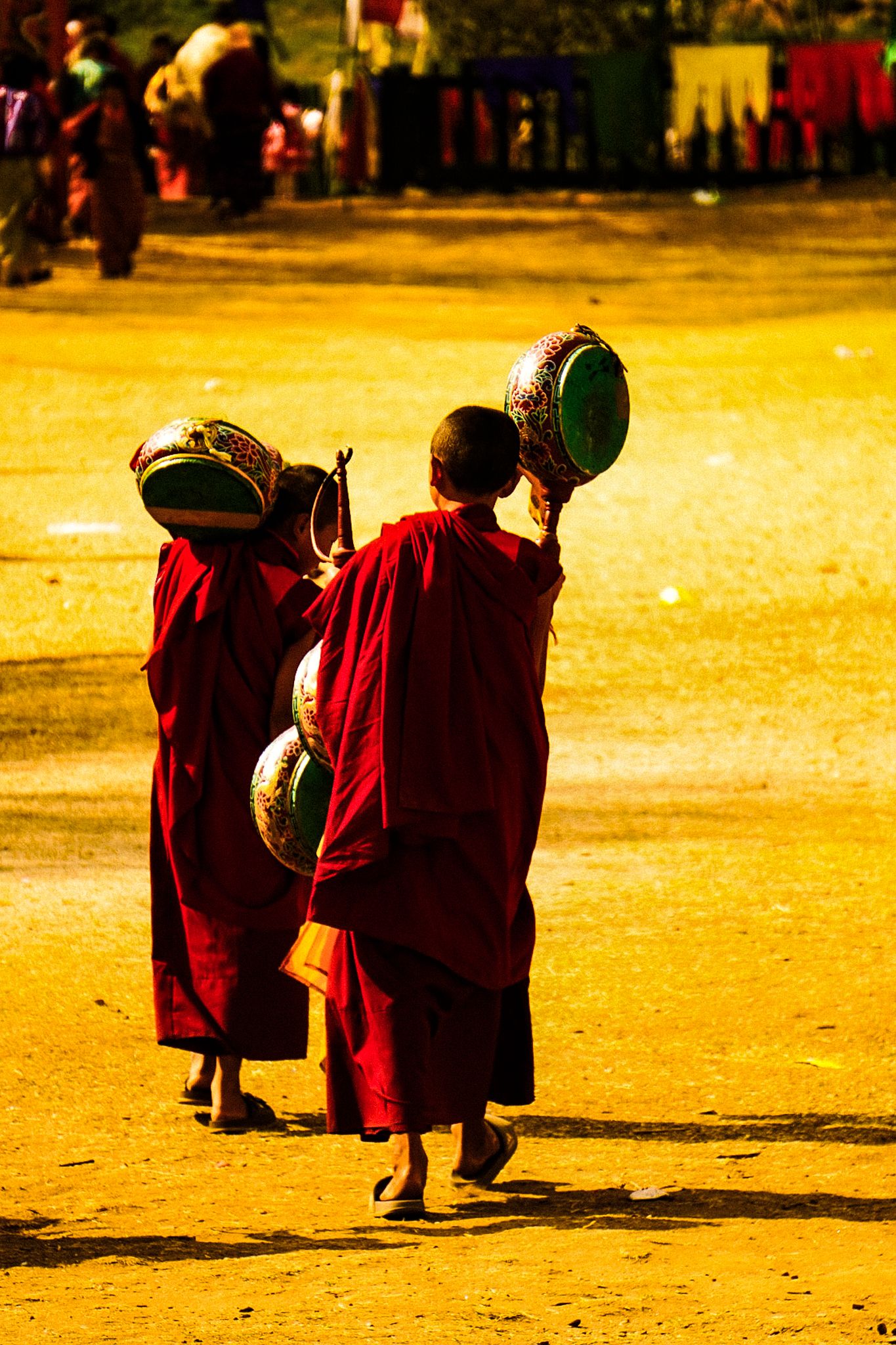 Two young Buddhist monks in red robes carrying ornate drums walking across golden sandy terrain.