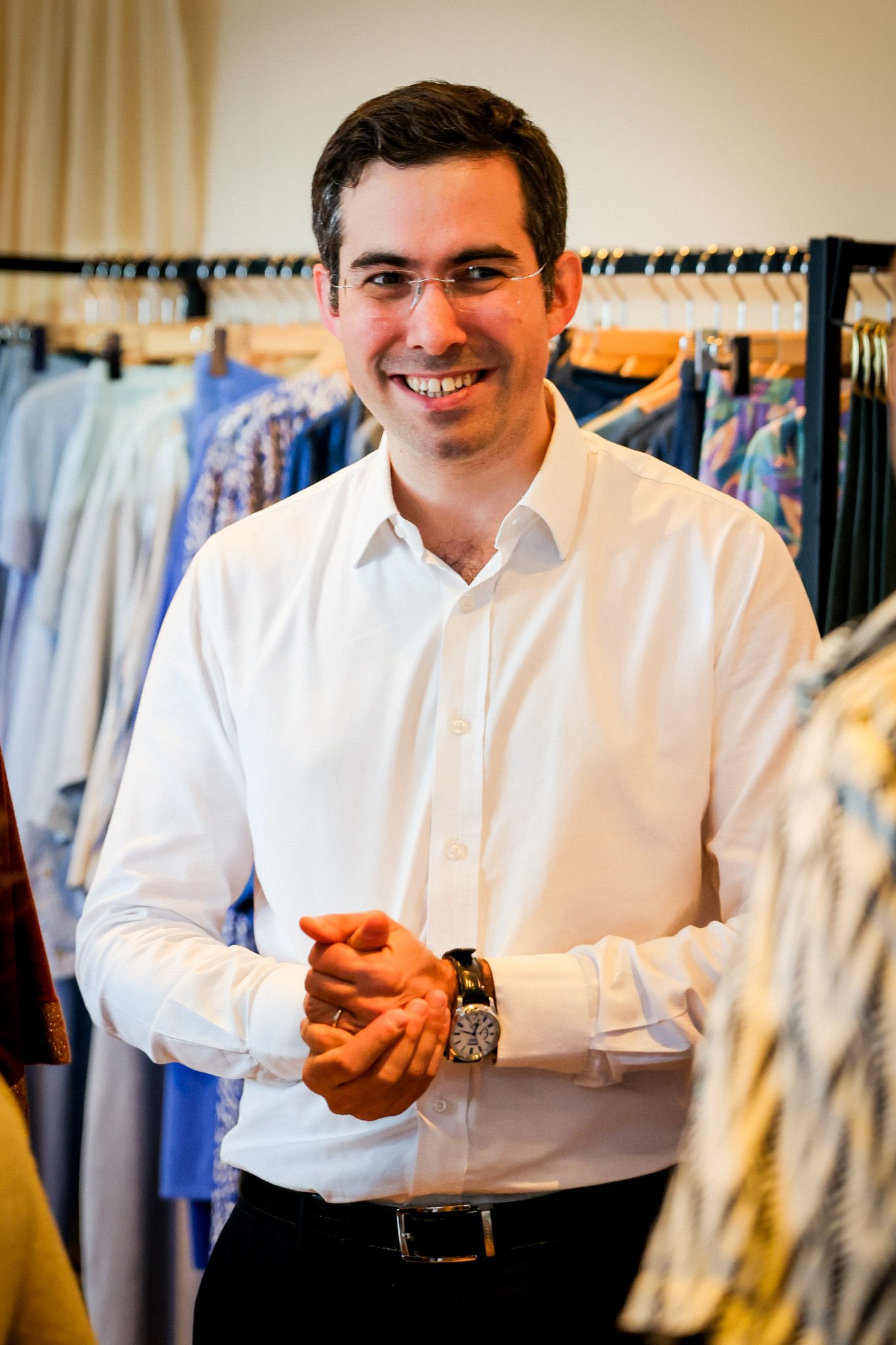 A smiling man in a white dress shirt stands confidently among clothing racks in a boutique store.