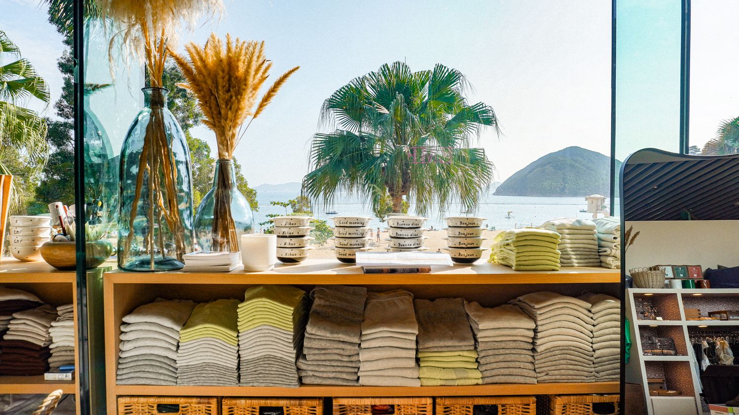 Wooden shelves displaying stacked colorful linens and ceramic bowls with tropical beach view through large window.