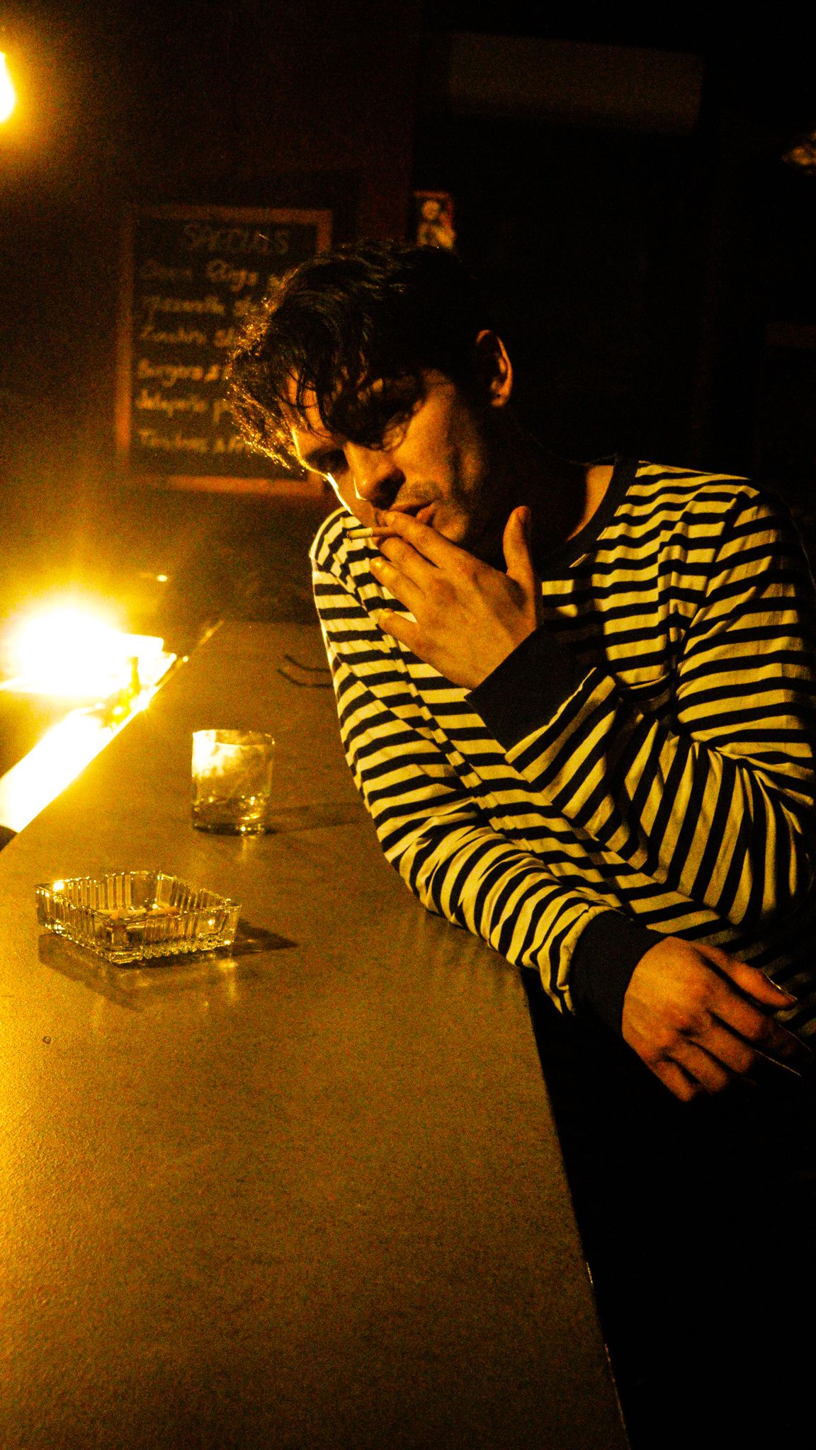 Young man in striped shirt rests his head on his hand at a dimly lit bar counter.