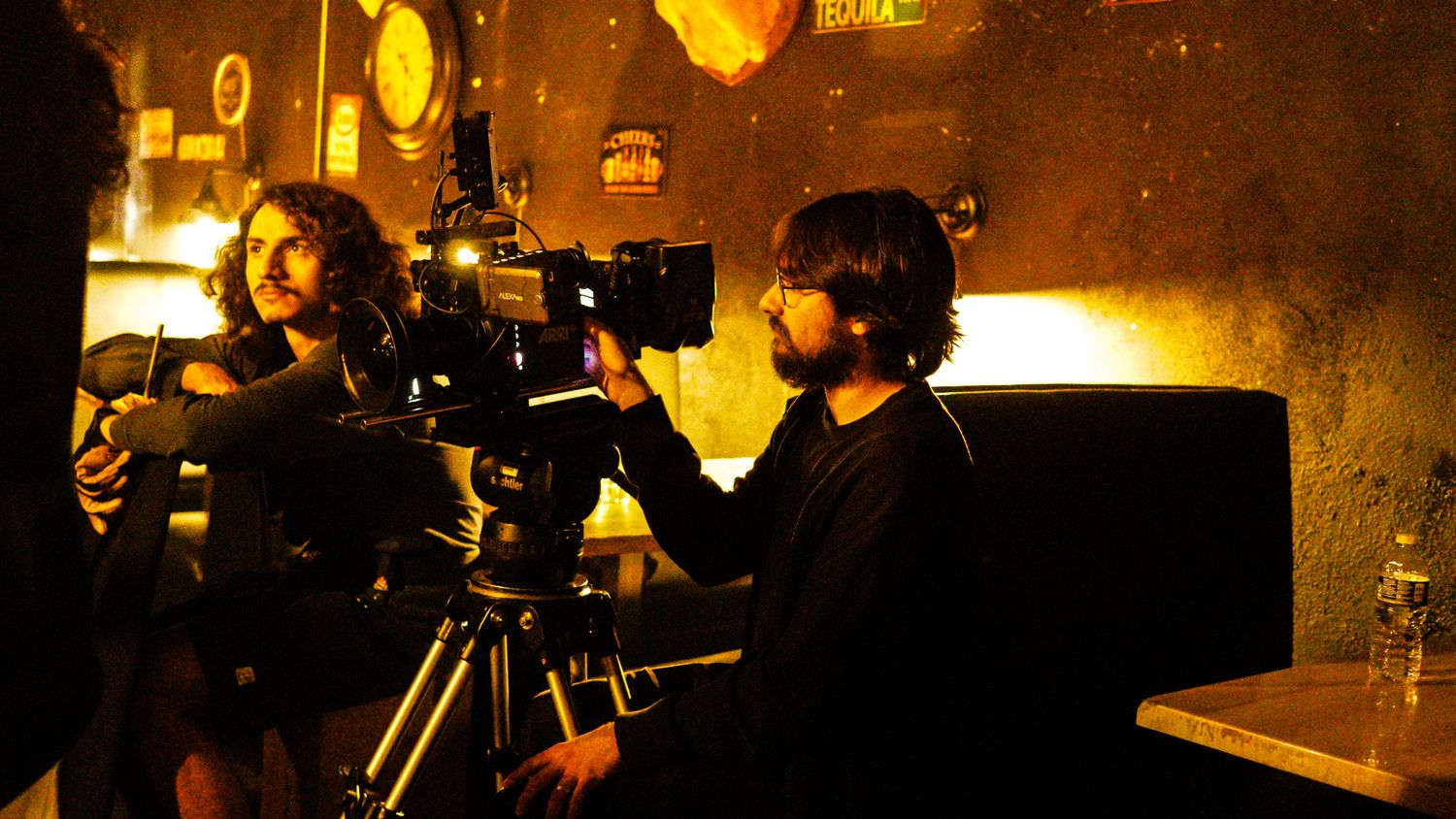 Film crew operating a cinema camera on a tripod in a dimly lit bar set with warm golden lighting.