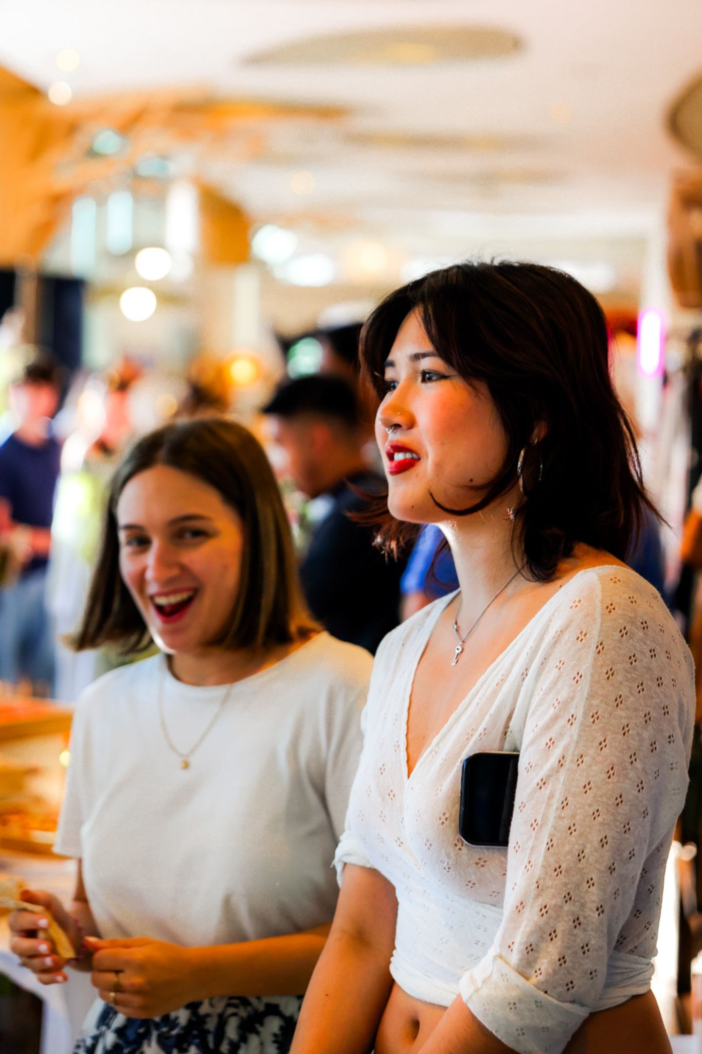 Two young women smiling and laughing together in a busy, warmly lit retail or event space.