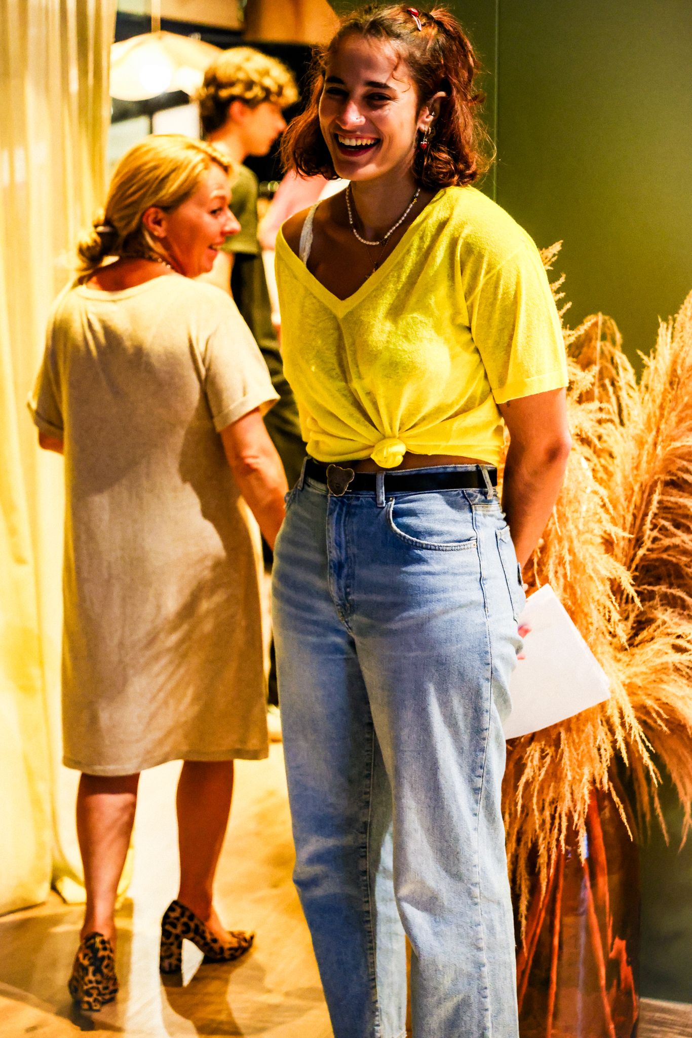 Smiling woman in yellow top and wide-leg jeans laughing in a warmly lit boutique store interior.
