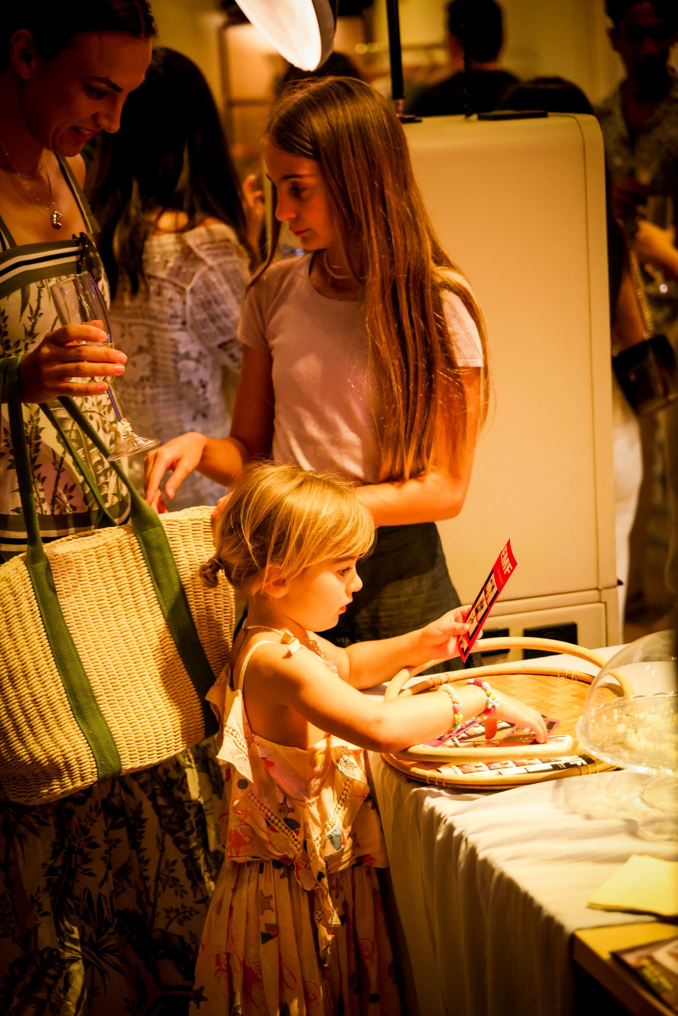 A young girl browses items on a display table at a boutique event while adults look on nearby.