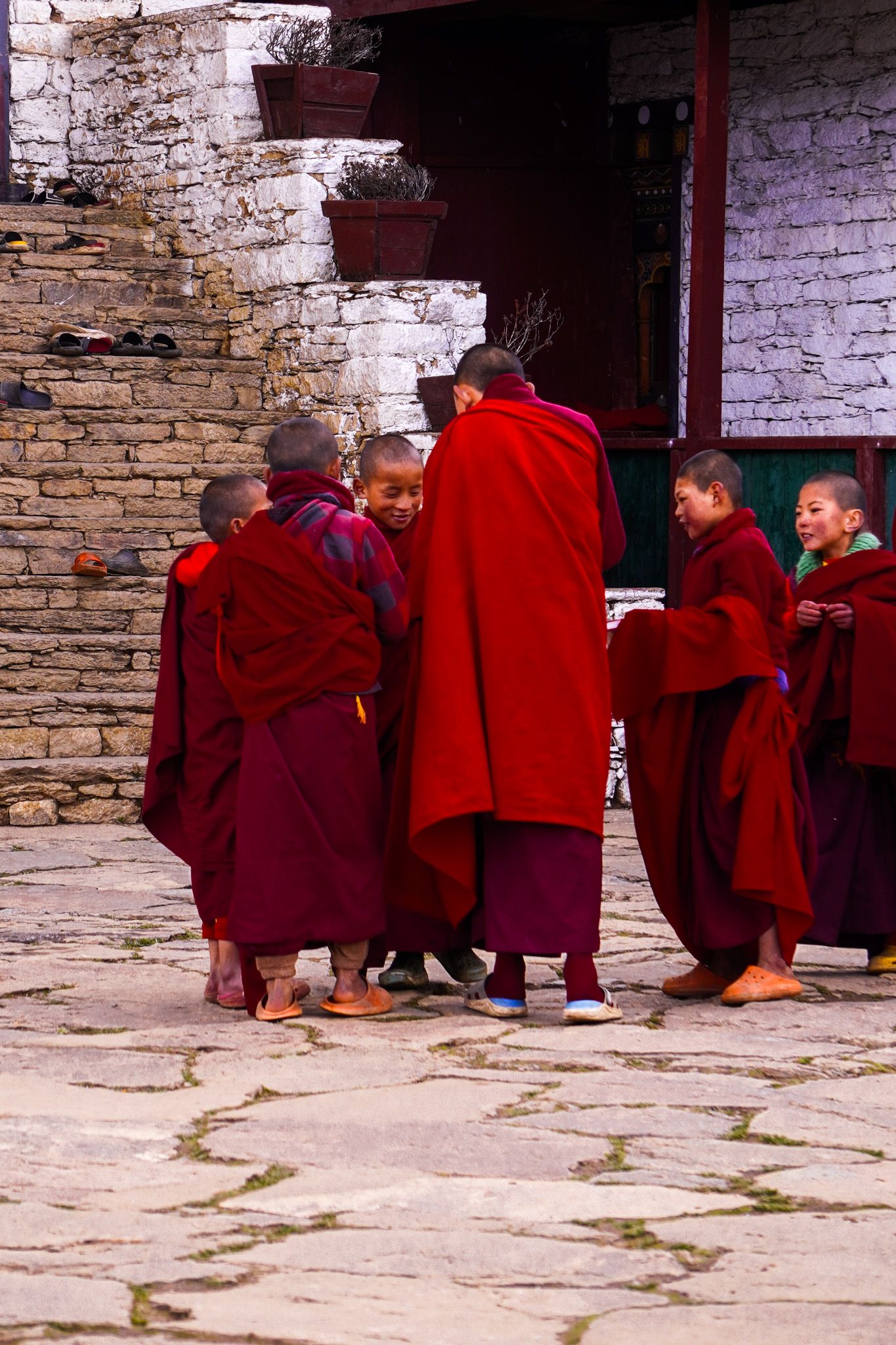 Young novice monks in red robes gather and chat in a stone courtyard of a Bhutanese monastery.