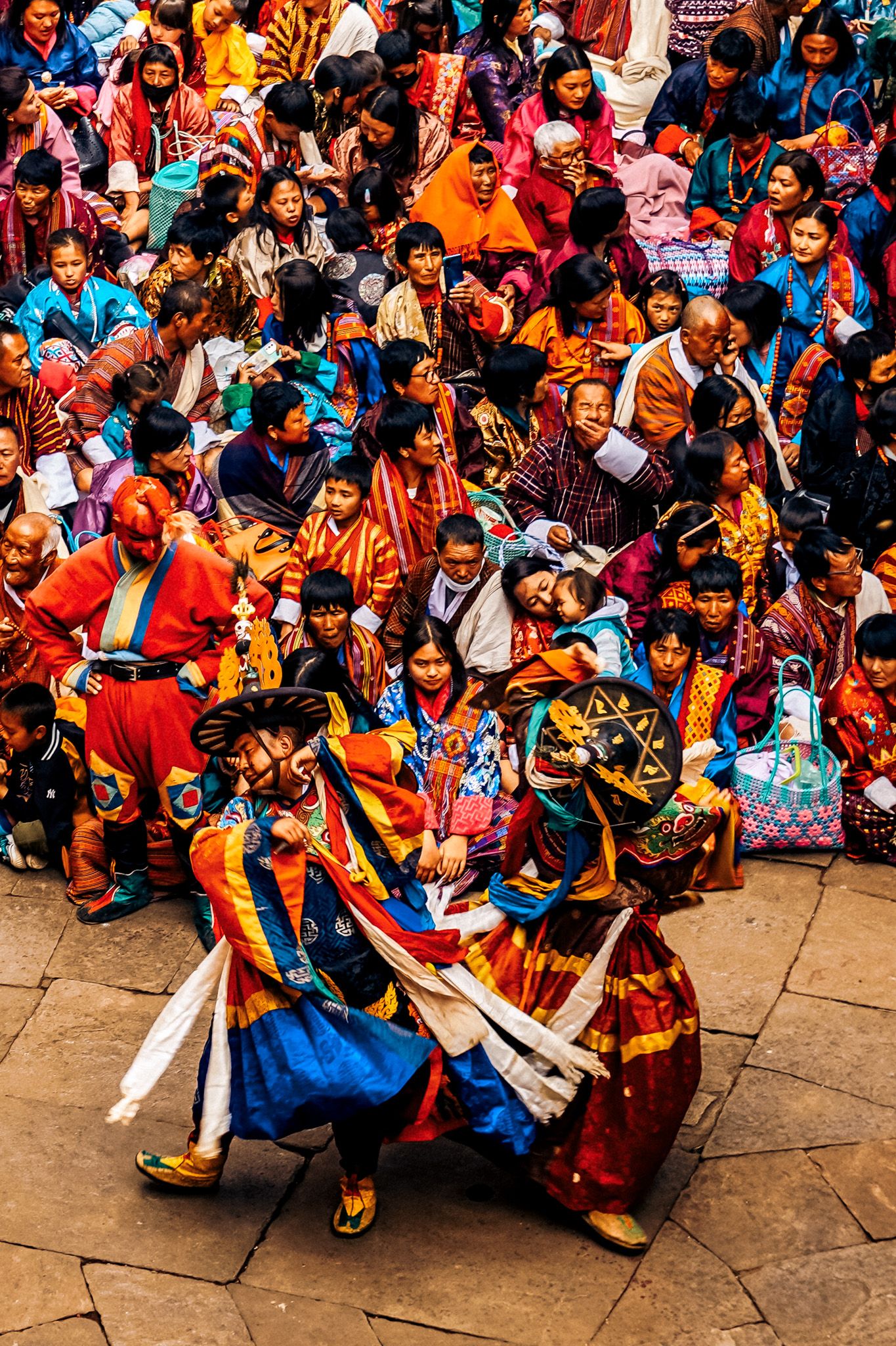 Bhutanese masked Cham dancers in vibrant silk costumes perform traditional sacred dance before a densely packed festival crowd.