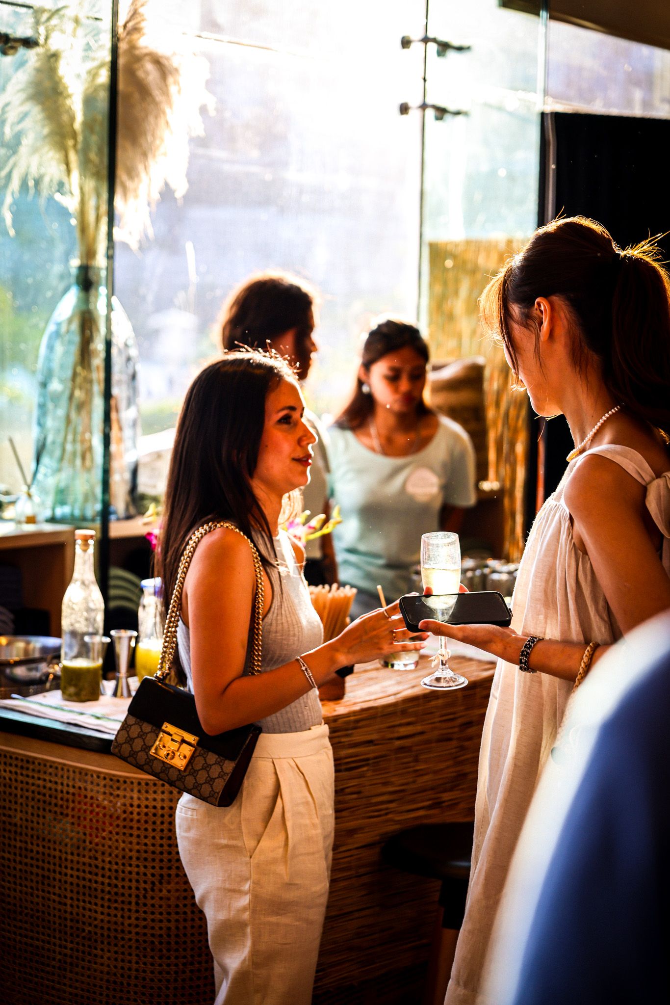 Two women in white outfits conversing at a bar counter with drinks in a warmly lit venue.