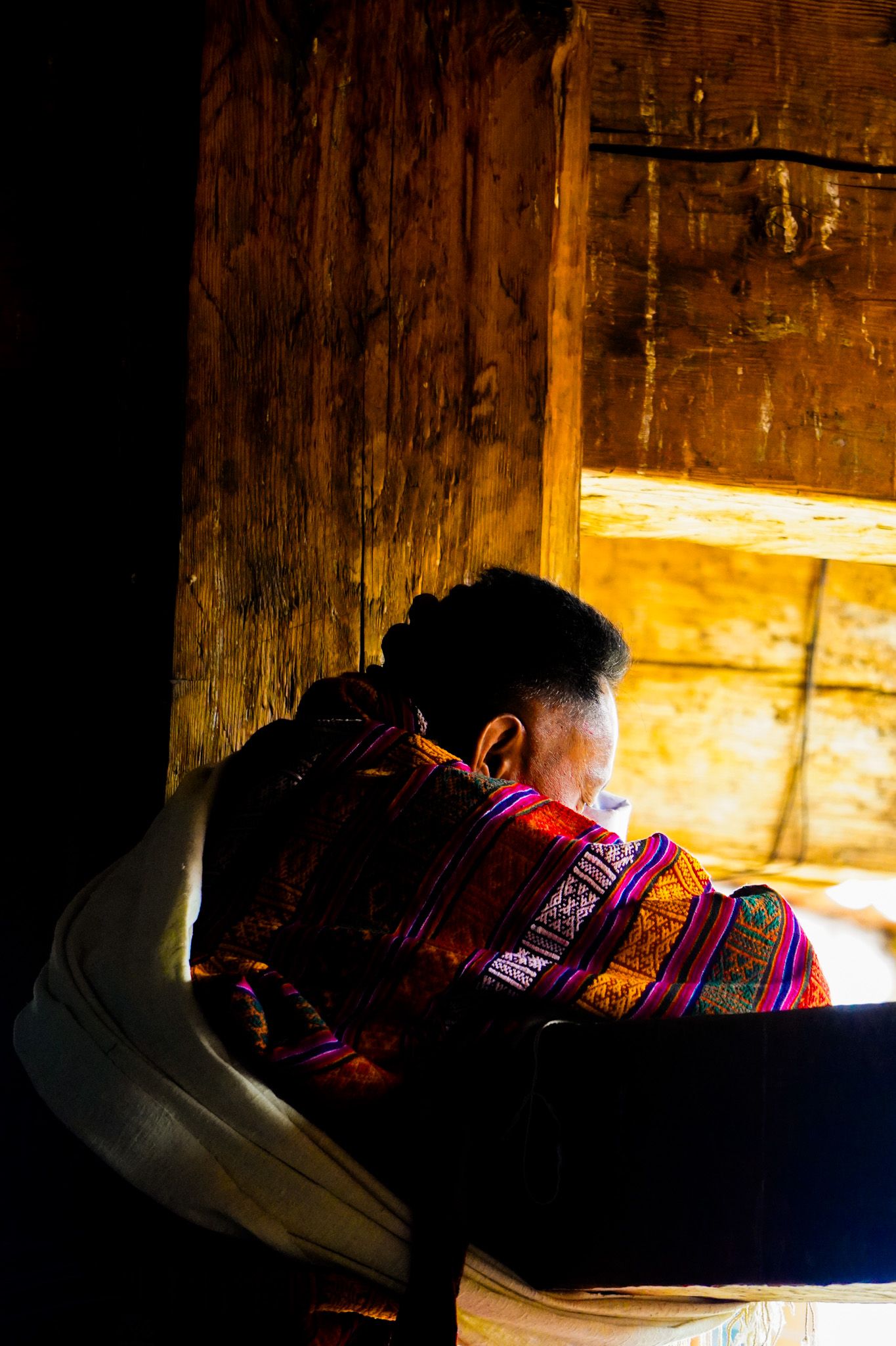 Elderly Bhutanese woman wearing vibrant traditional textile wrap sits by rustic wooden window.