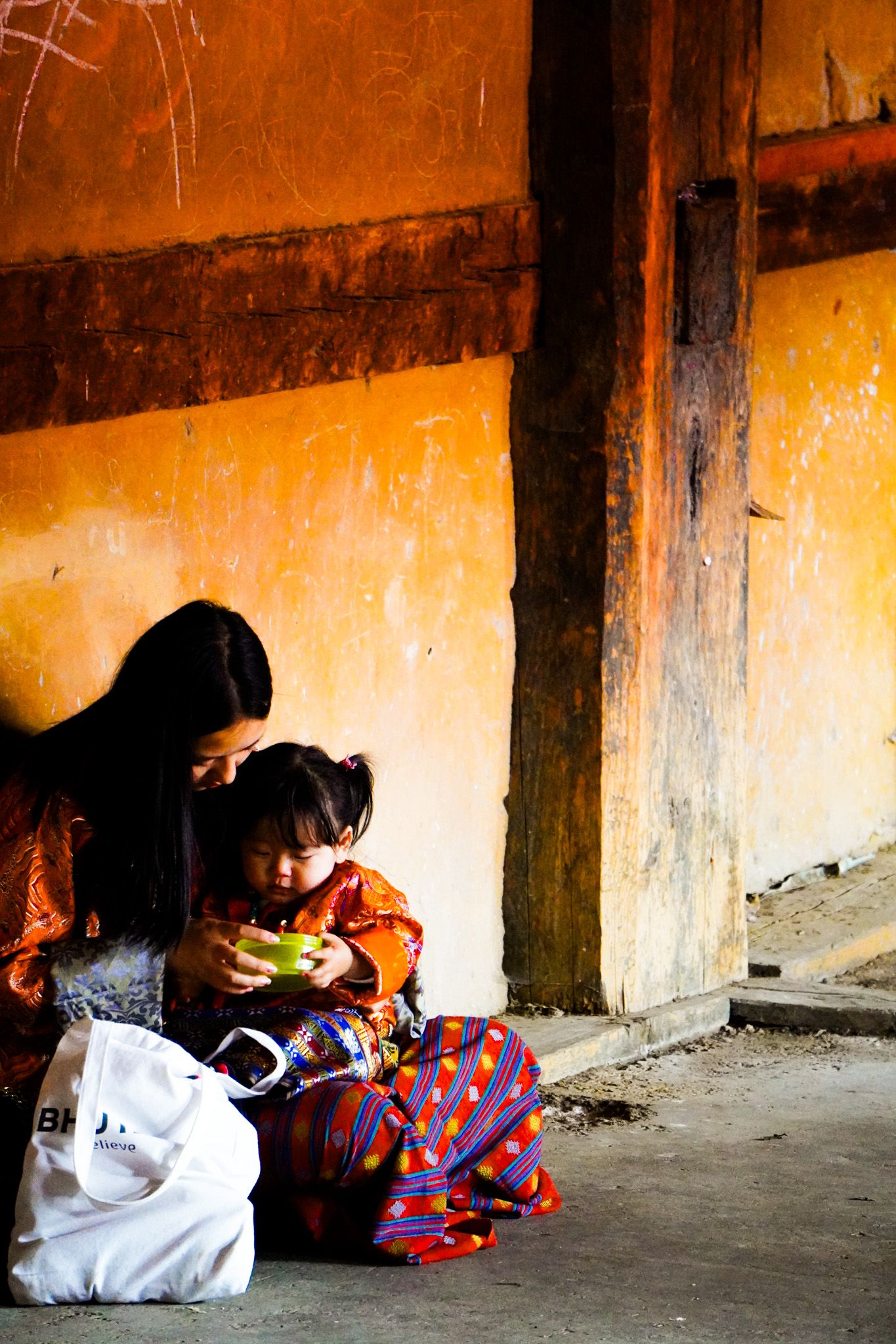 Bhutanese mother and young child in traditional dress sit against orange wall looking at phone.