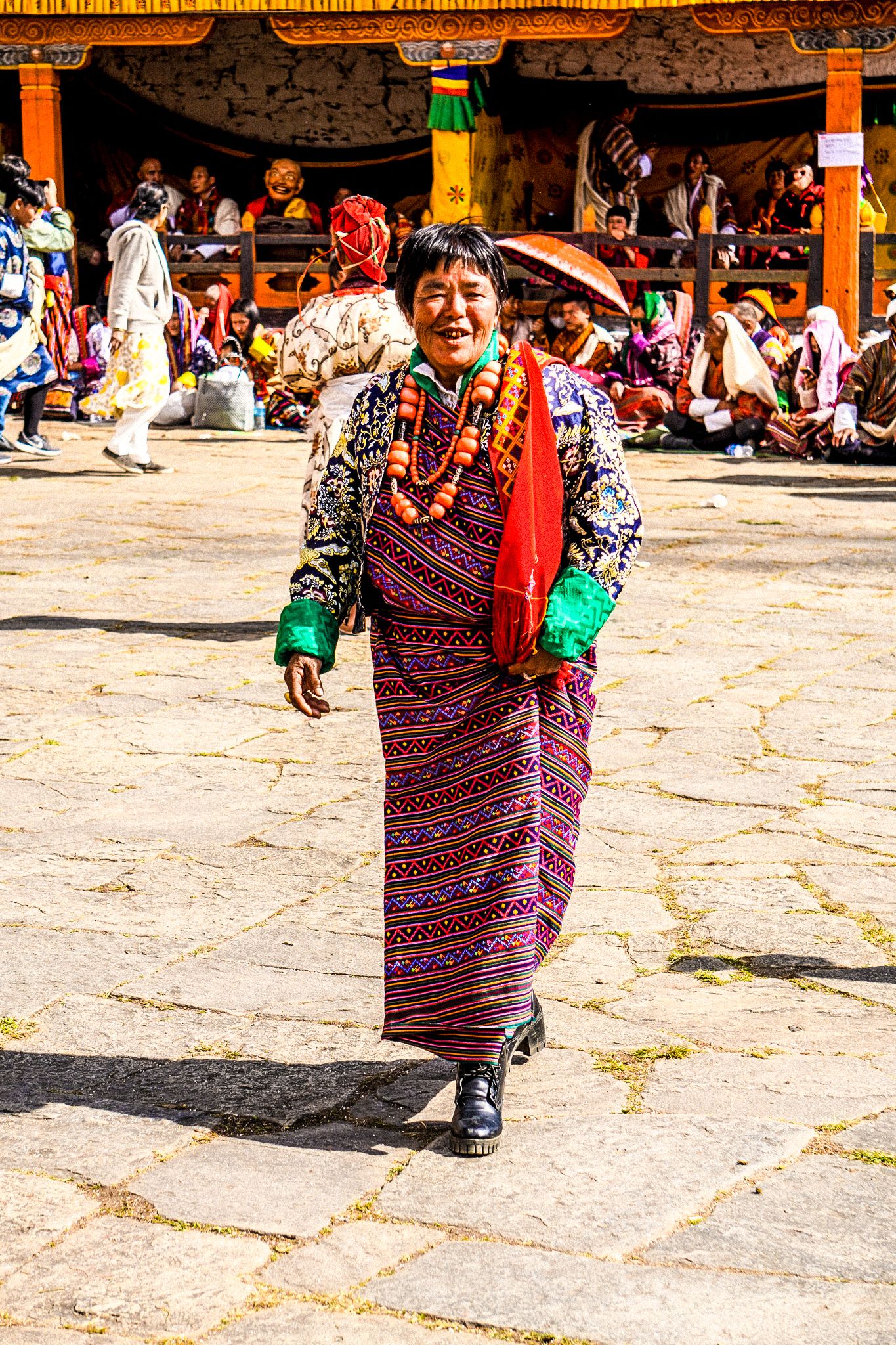 Smiling Bhutanese woman in vibrant traditional kira and beaded necklace walking in monastery courtyard.