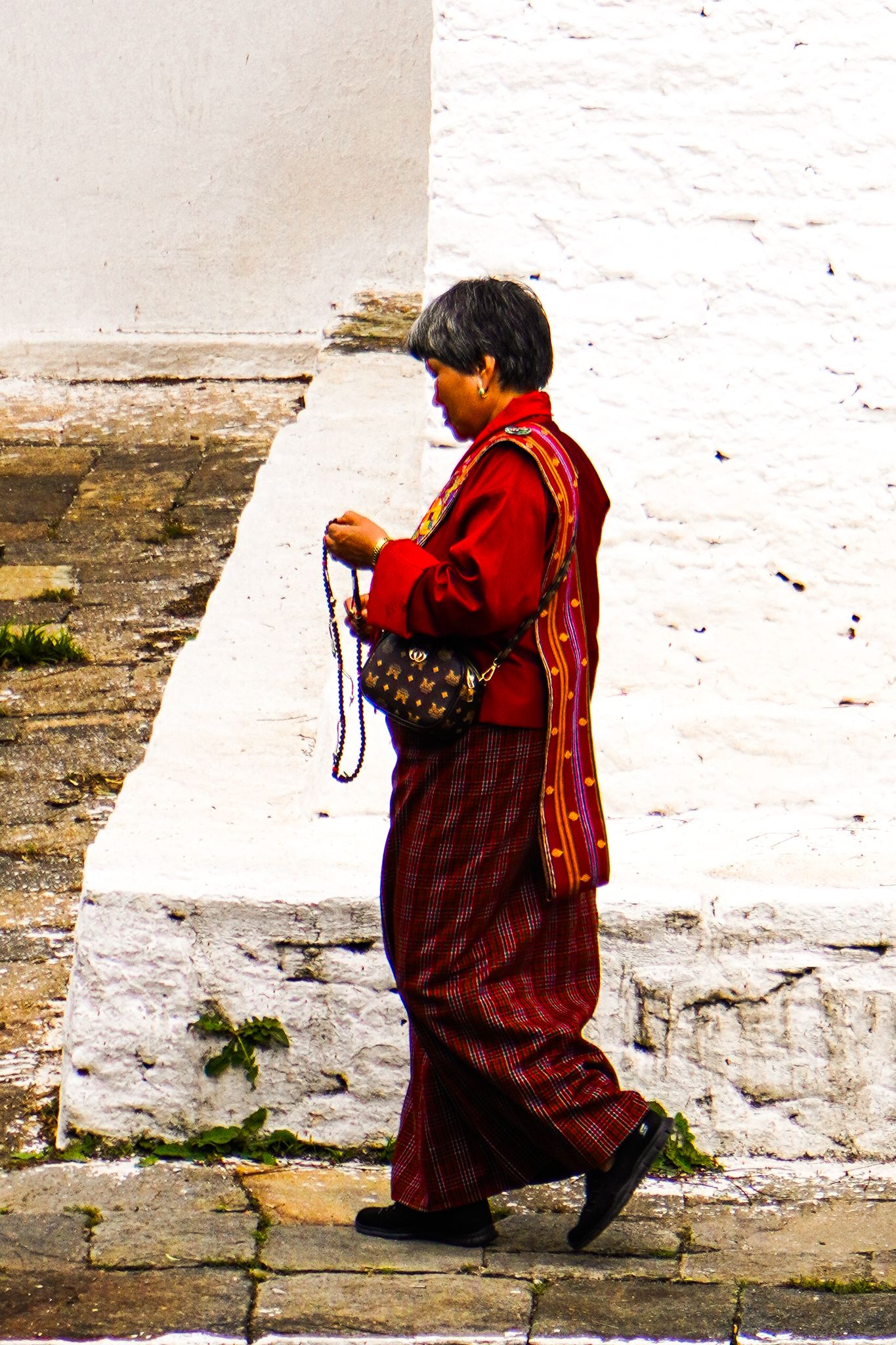 A Bhutanese woman in traditional red kira walks while holding prayer beads near a white monastery wall.