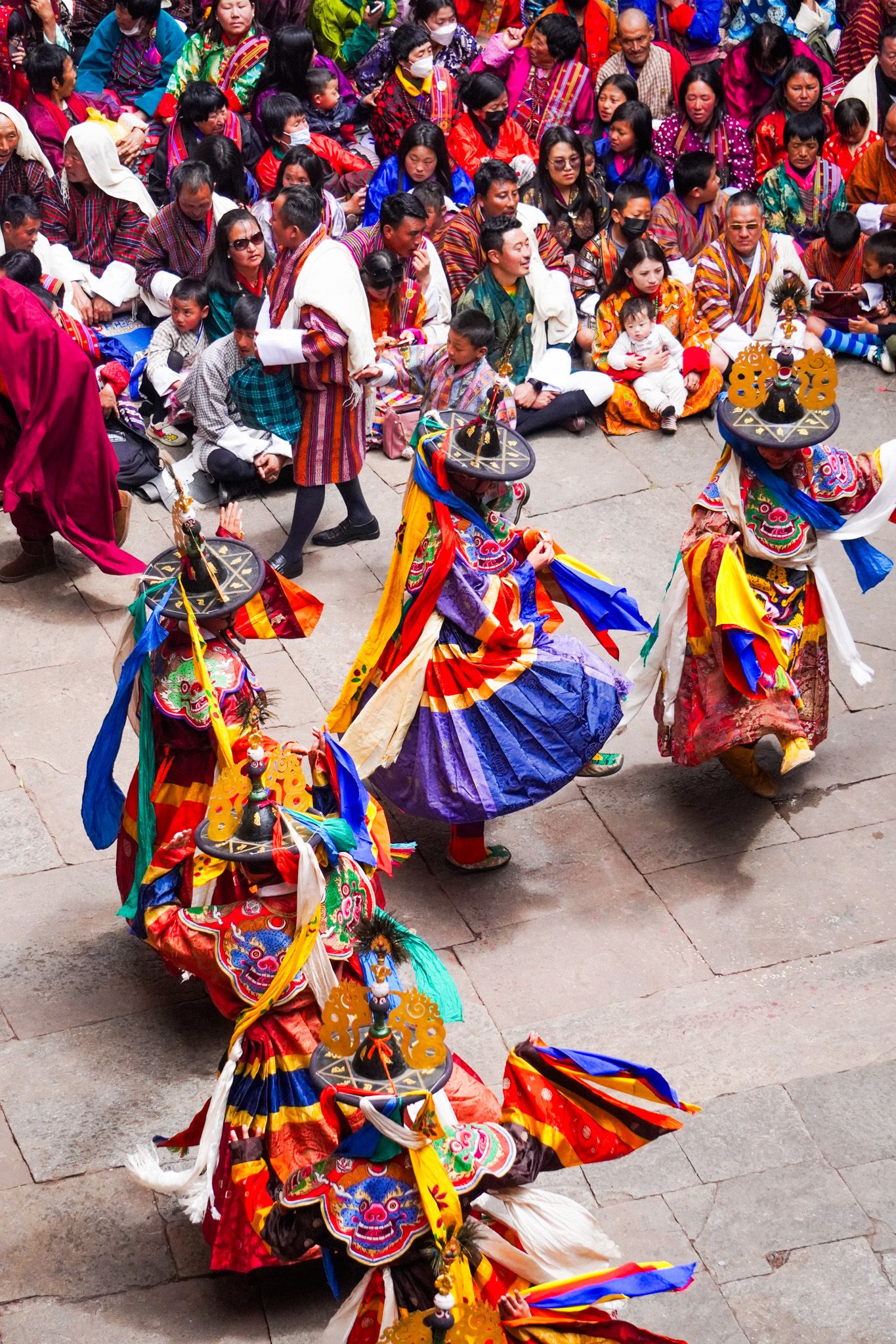 Four ornately costumed Bhutanese Cham dancers wearing decorative masks perform traditional sacred Tsechu festival dance for spectators.
