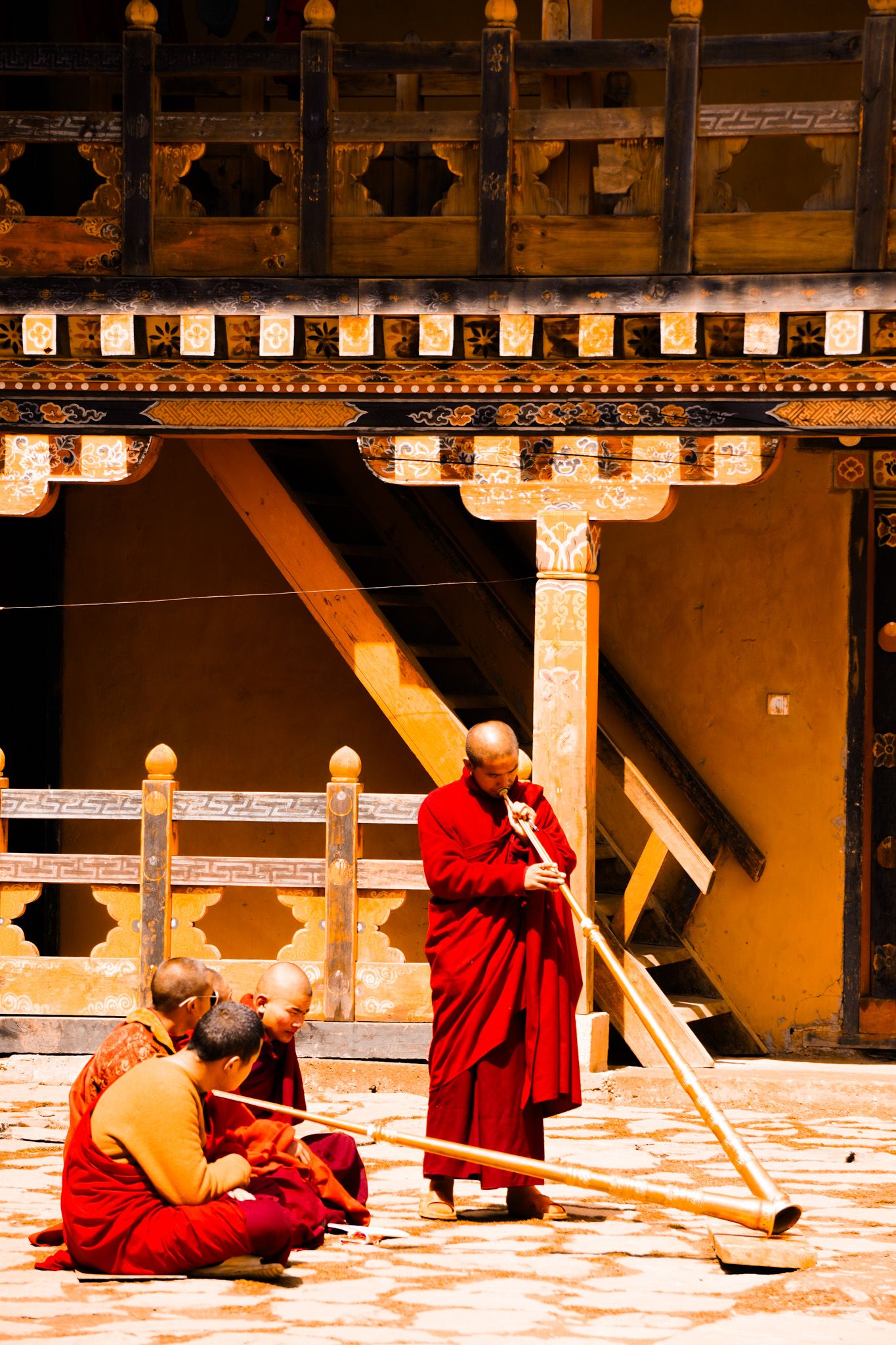 Buddhist monks in red robes play traditional long horn instruments in an ornate Bhutanese dzong courtyard.