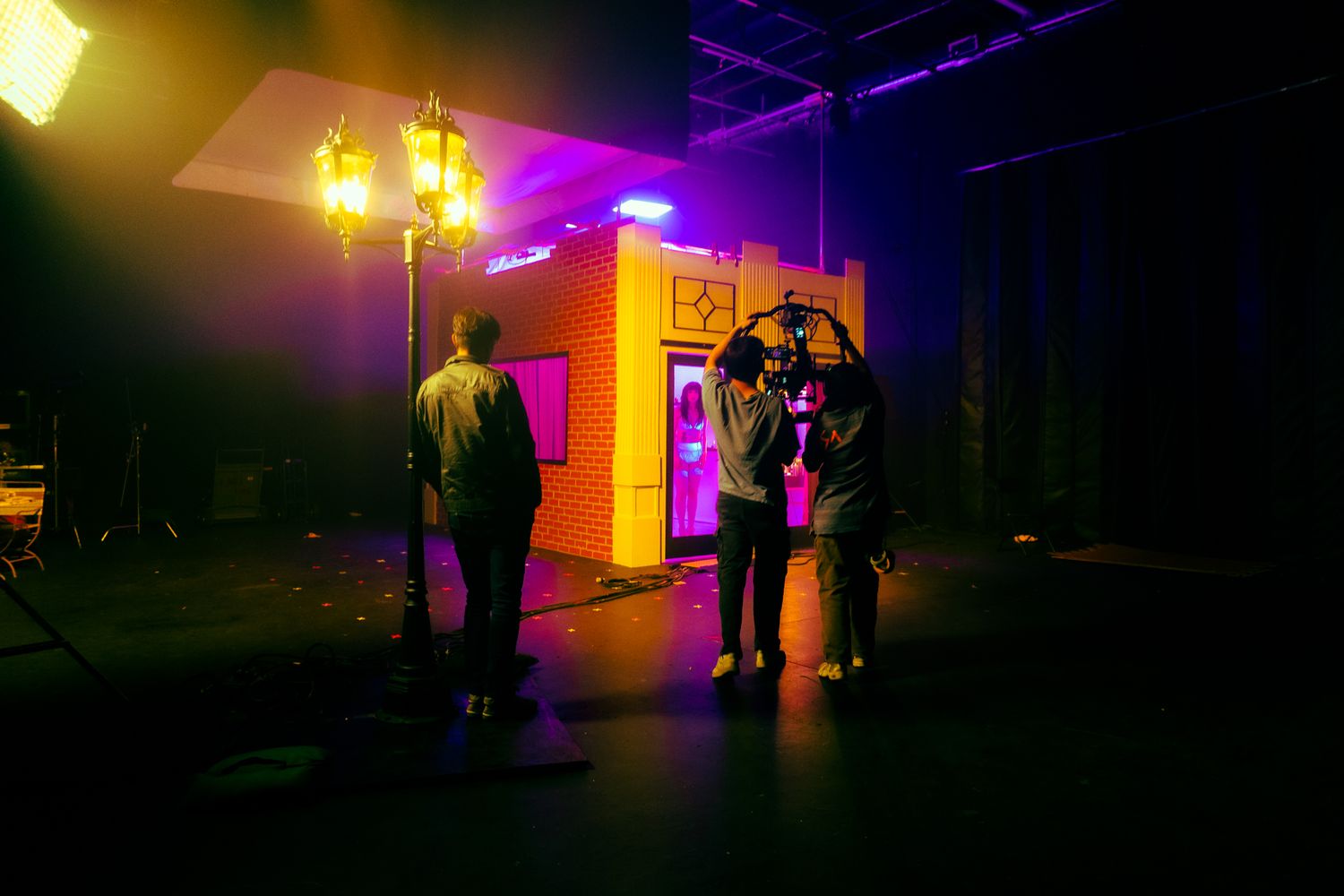 Small film crew shoots a scene near a colorful neon-lit brick storefront prop on a dark studio set.
