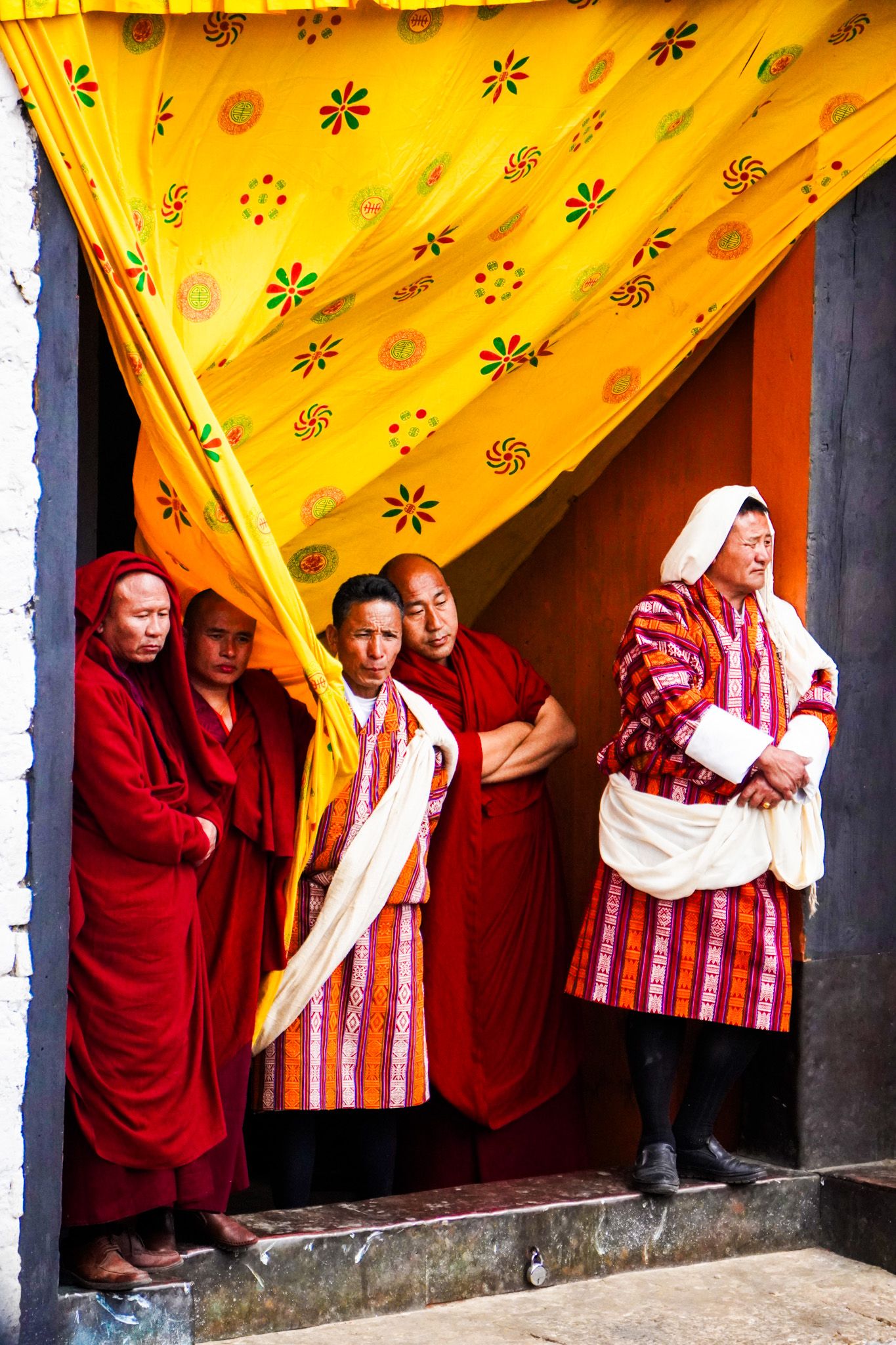 Buddhist monks and laypeople in traditional dress stand beneath a yellow ceremonial canopy in Bhutan.