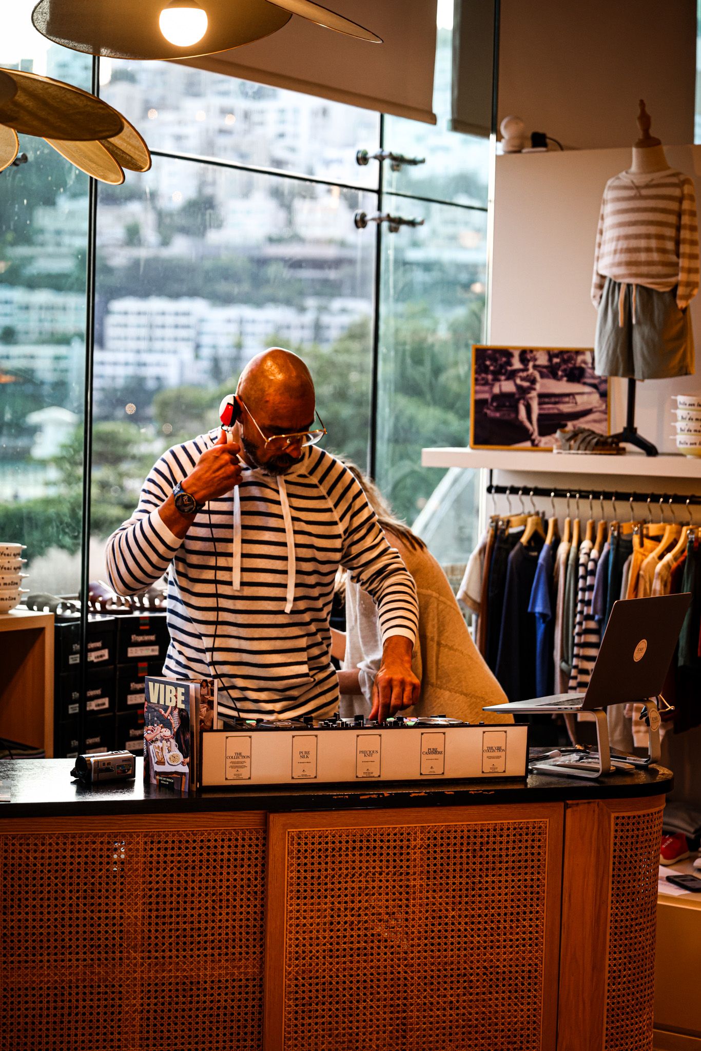 Bald man in striped hoodie talking on phone behind retail counter with laptop and clothing racks in background.