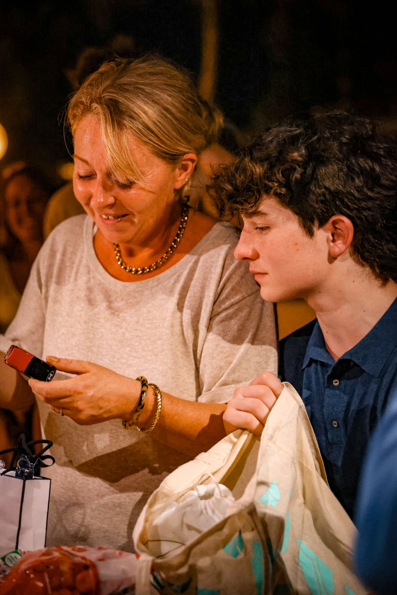 Blonde woman and teenage boy examining items together at an outdoor evening event.