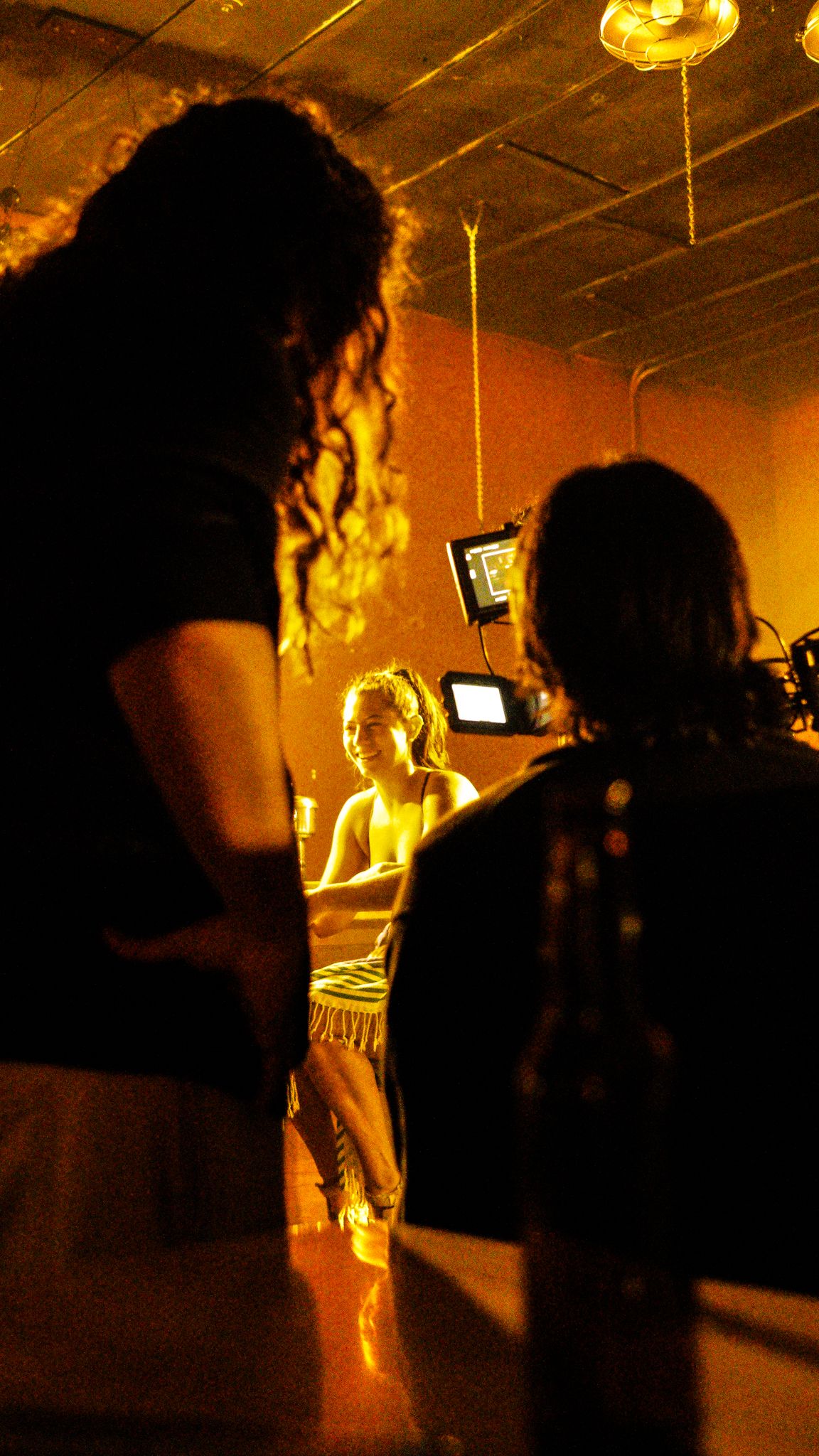 Film crew members observe an actor performing at a bar set bathed in warm amber lighting.