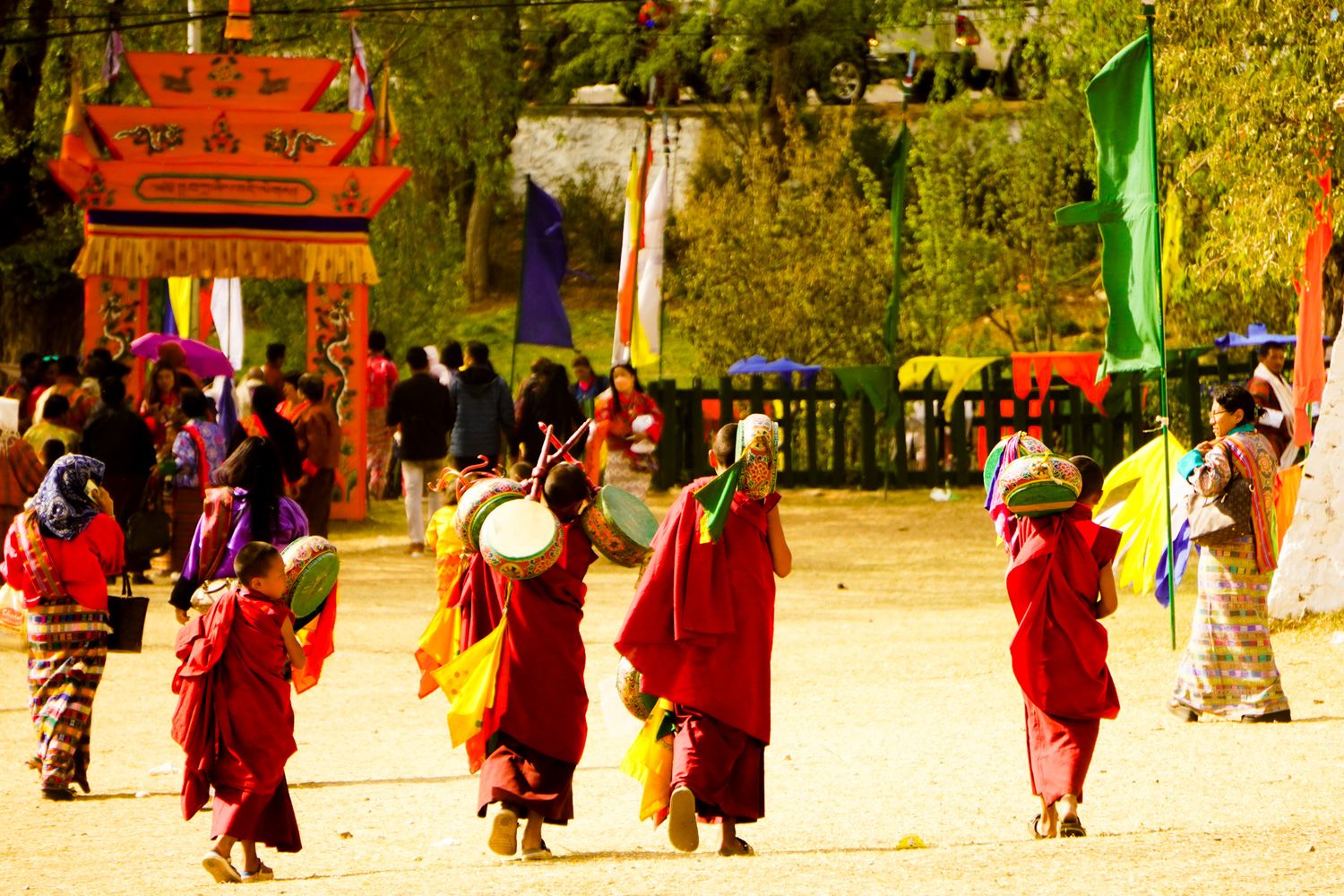 Group of Buddhist monks in red robes carrying drums walking through a colorful festival grounds.