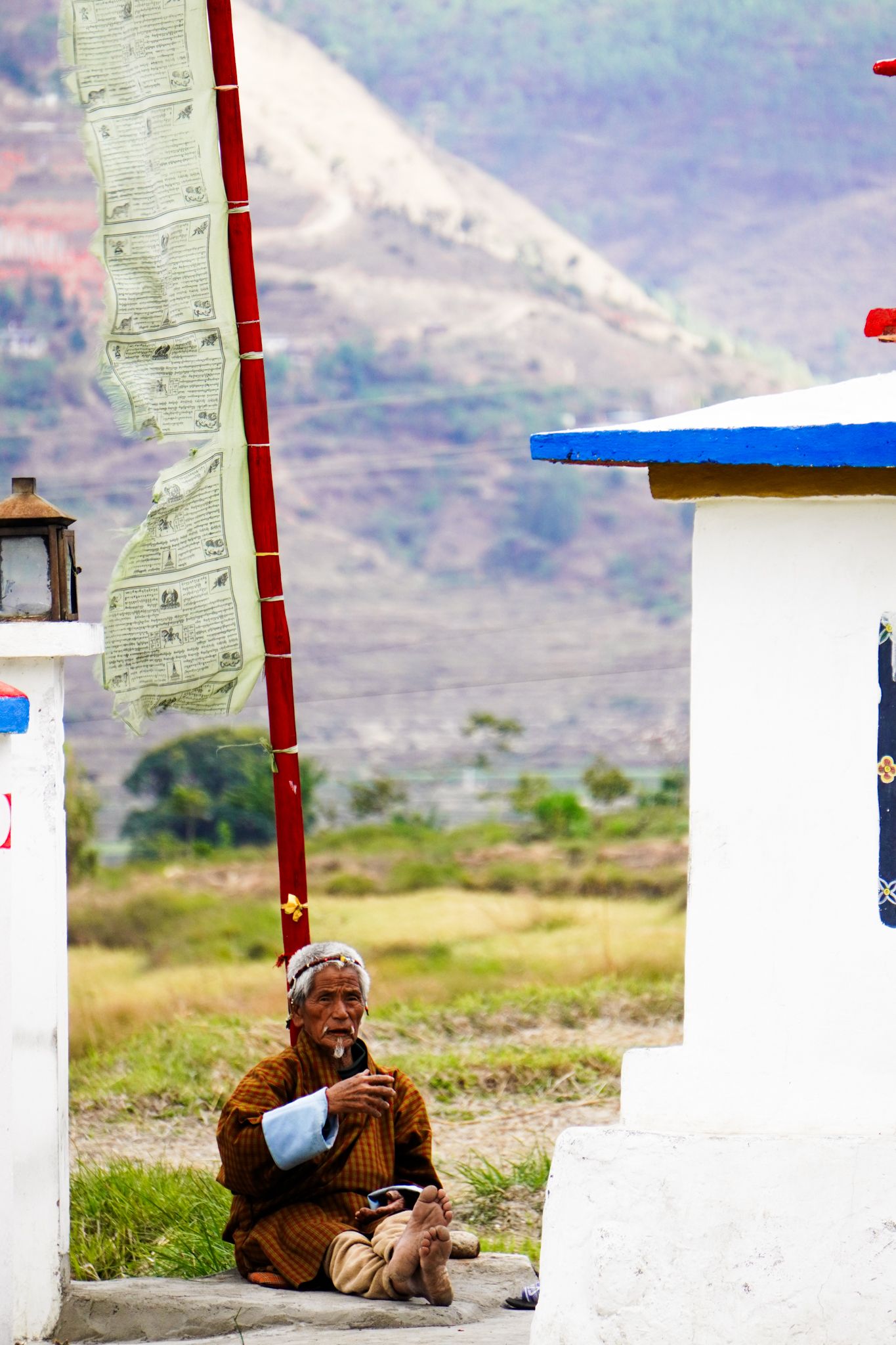 Elderly Bhutanese man in traditional robes sits beside prayer flag pole with mountain backdrop.