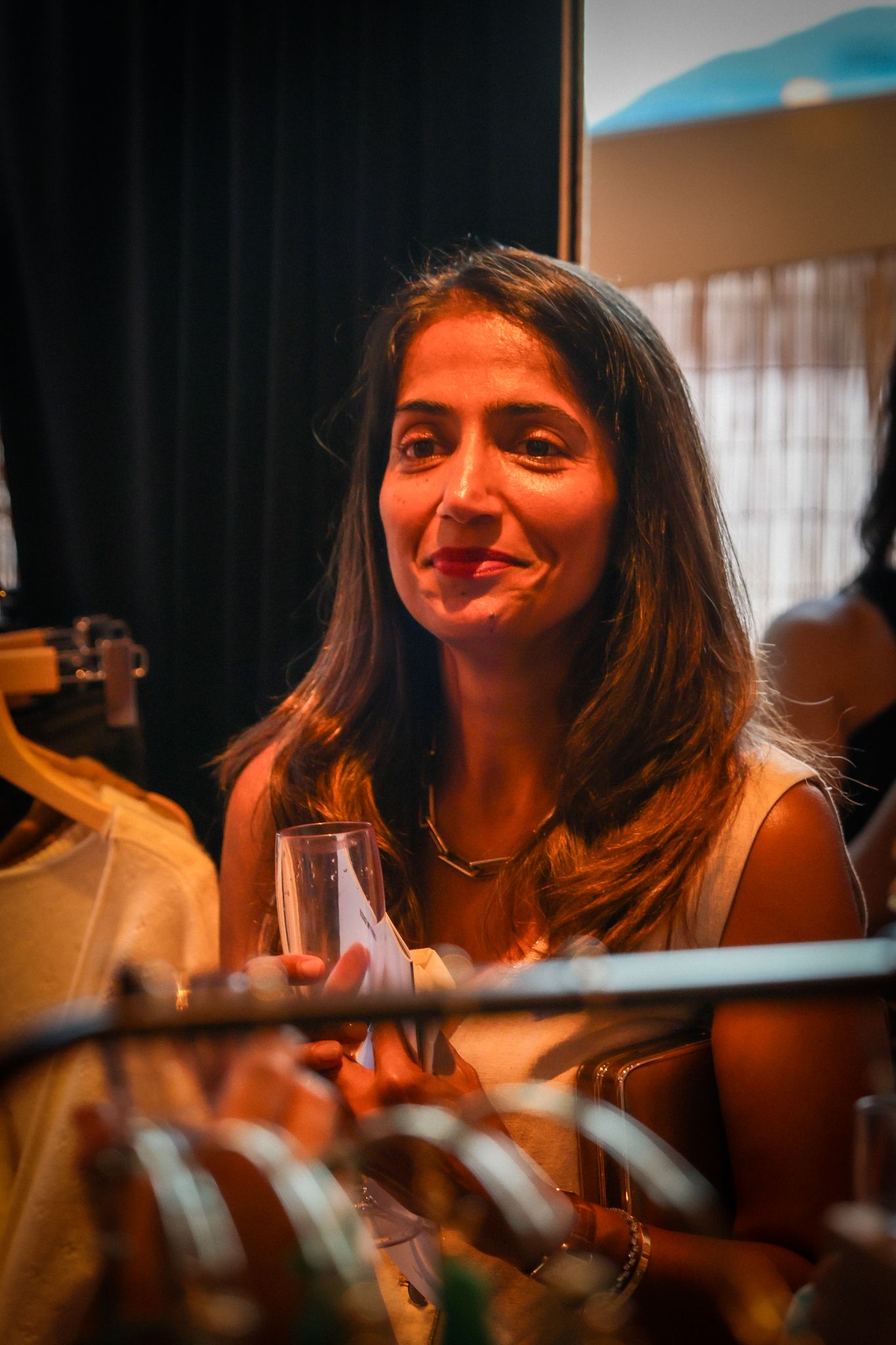 Woman with long hair smiling and holding a glass at an indoor shopping event.