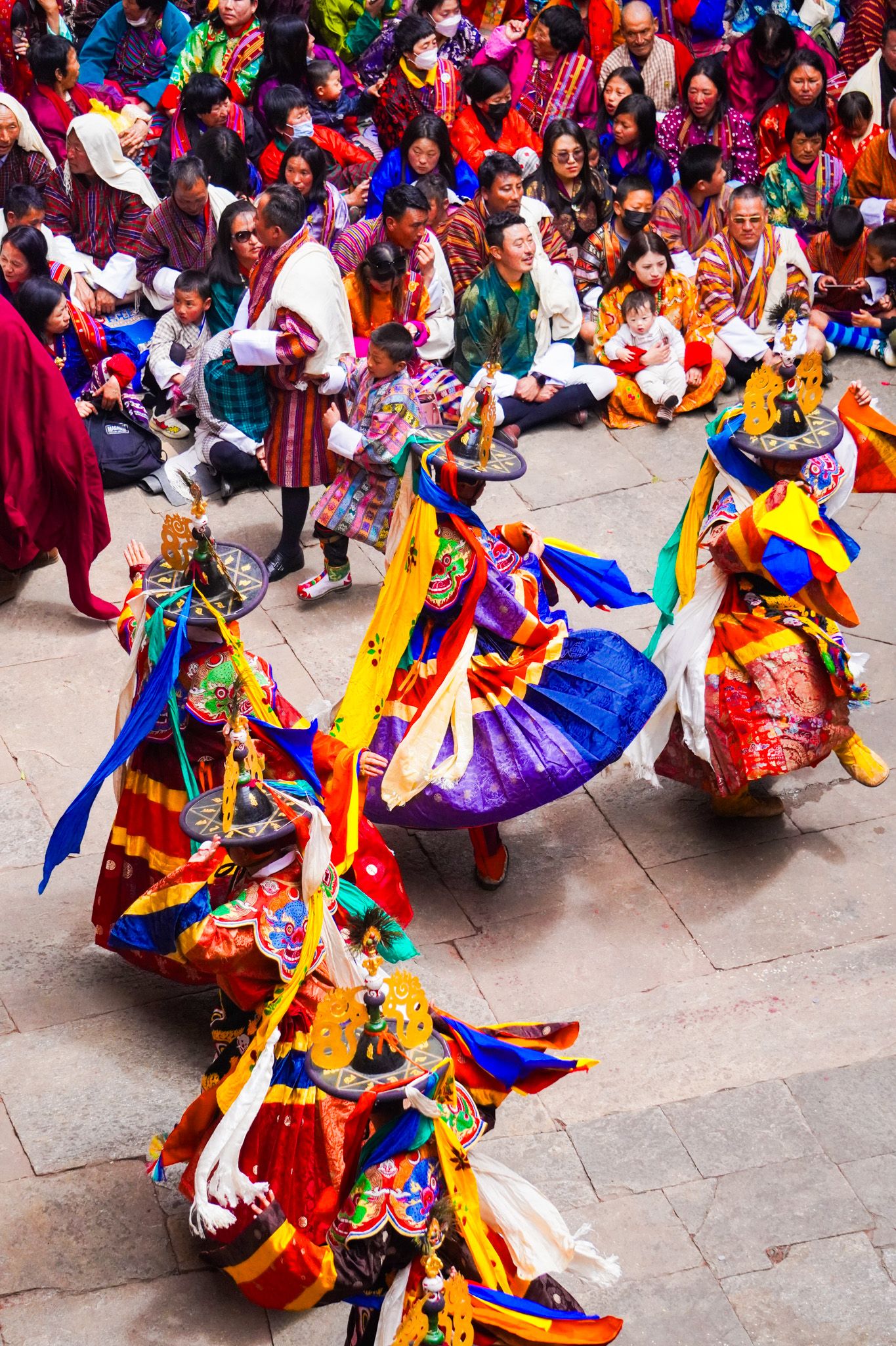Colorfully costumed Bhutanese Cham dancers spin in elaborate silk robes during a traditional sacred Tsechu festival performance.