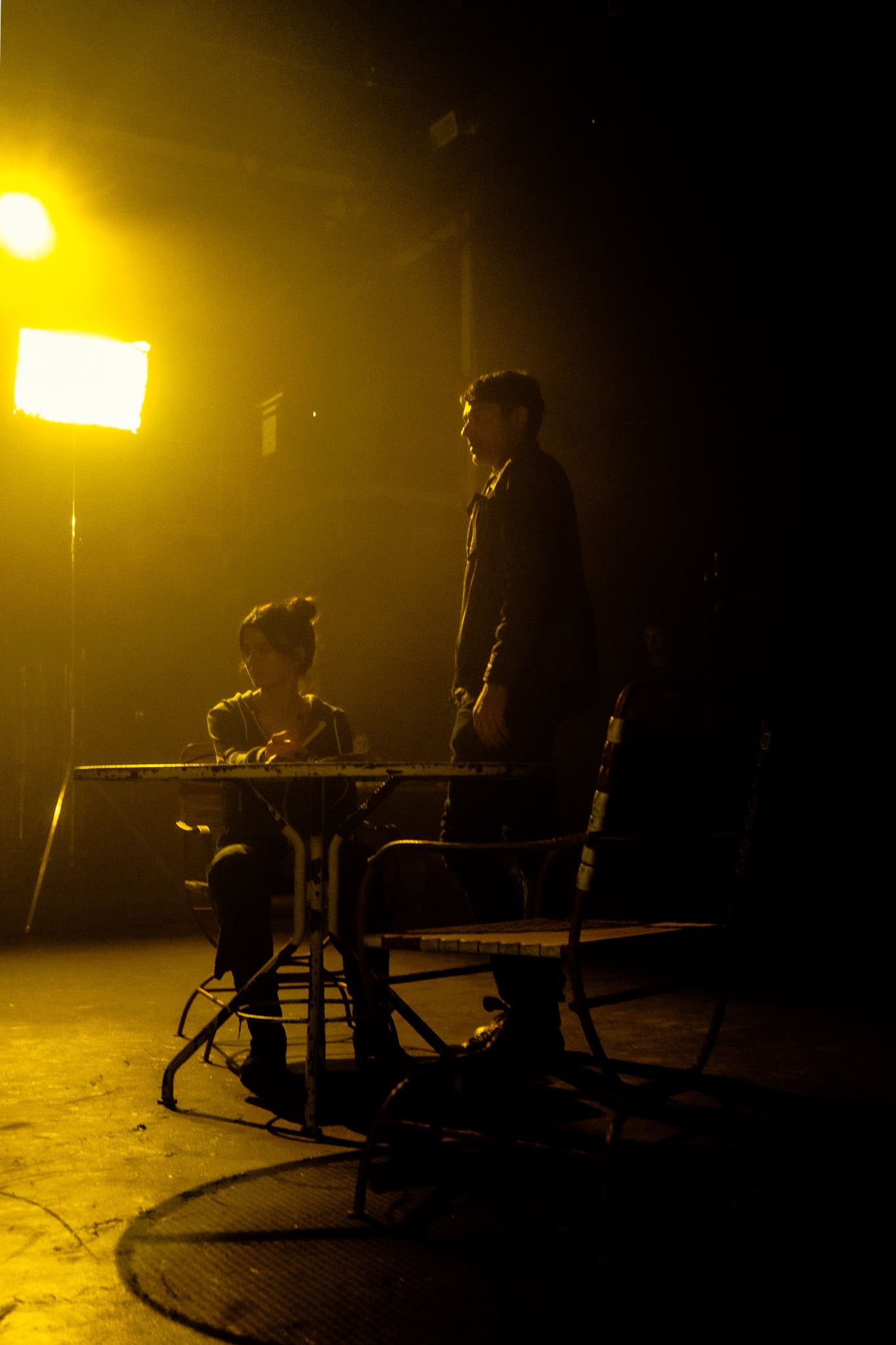 Two silhouetted figures at a table on a dark stage illuminated by bright yellow studio lighting equipment.
