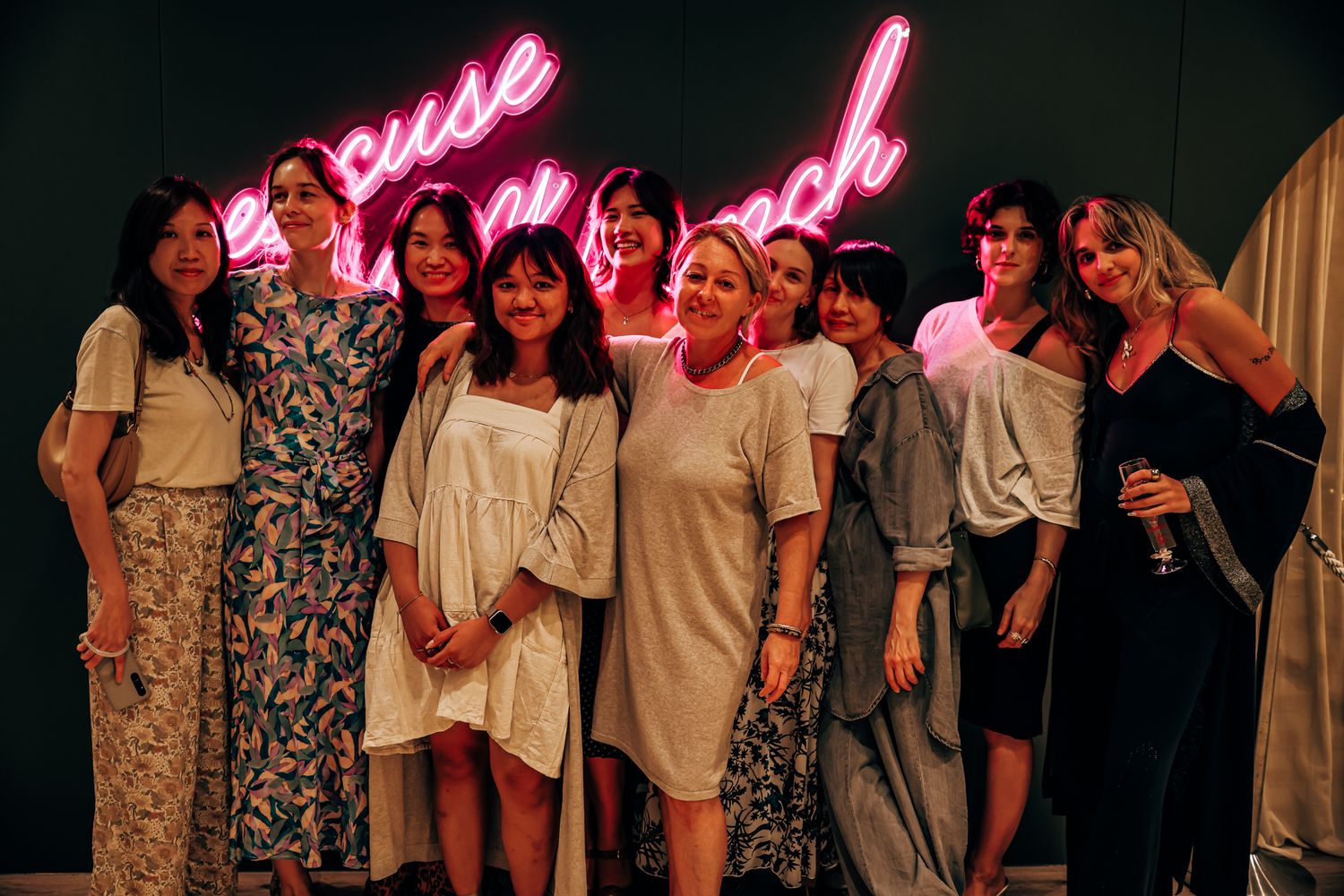 Smiling group of women gathered in front of a glowing pink neon sign at a party.