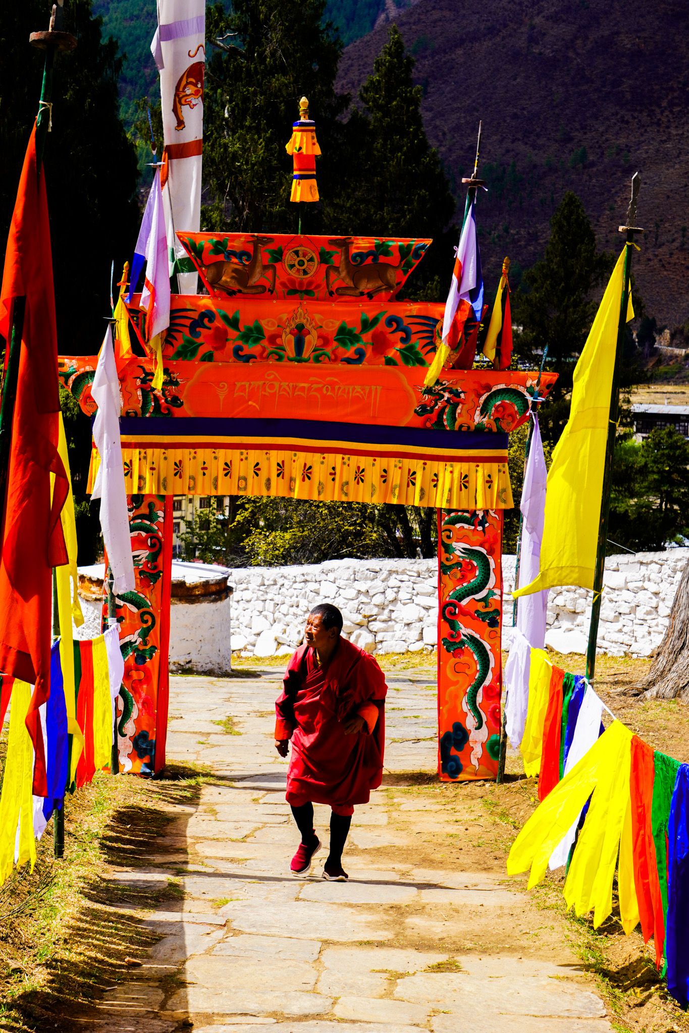 Buddhist monk in red robes walks through a decorated ceremonial gate with colorful prayer flags.