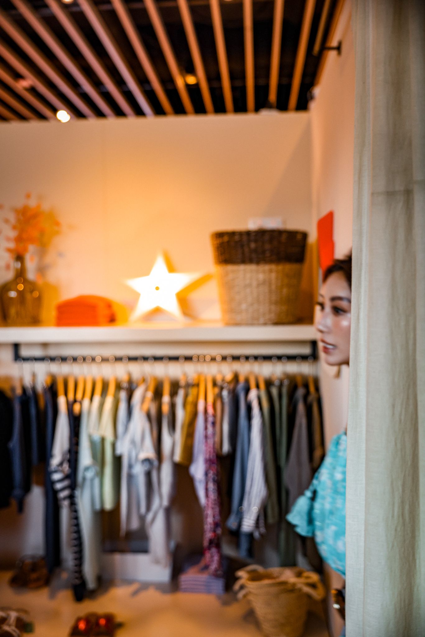 A woman peeks around a corner near a clothing rack in a warmly lit boutique with a star lamp.
