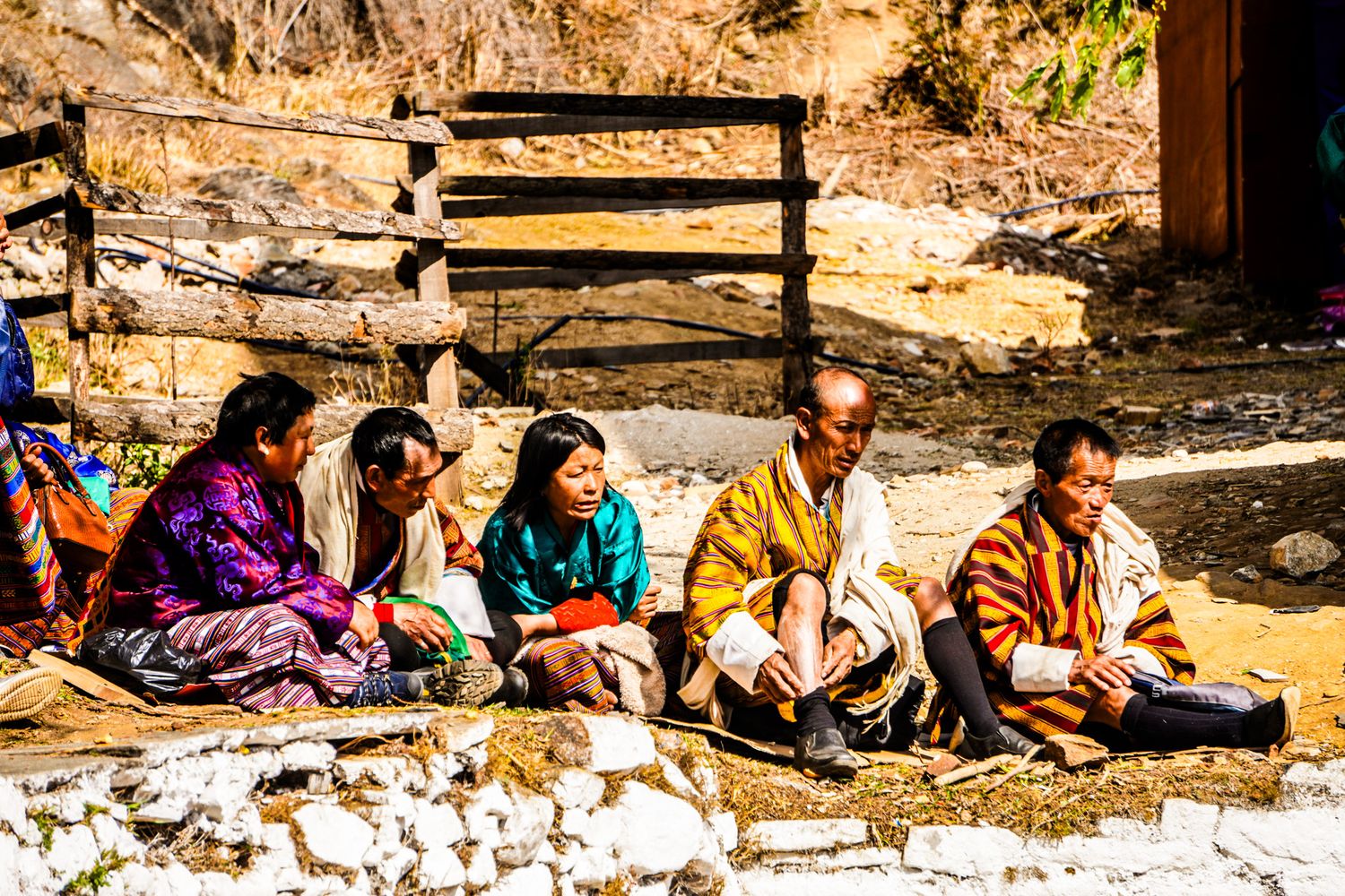 Group of Bhutanese people in traditional kira and gho attire sitting outdoors on the ground.