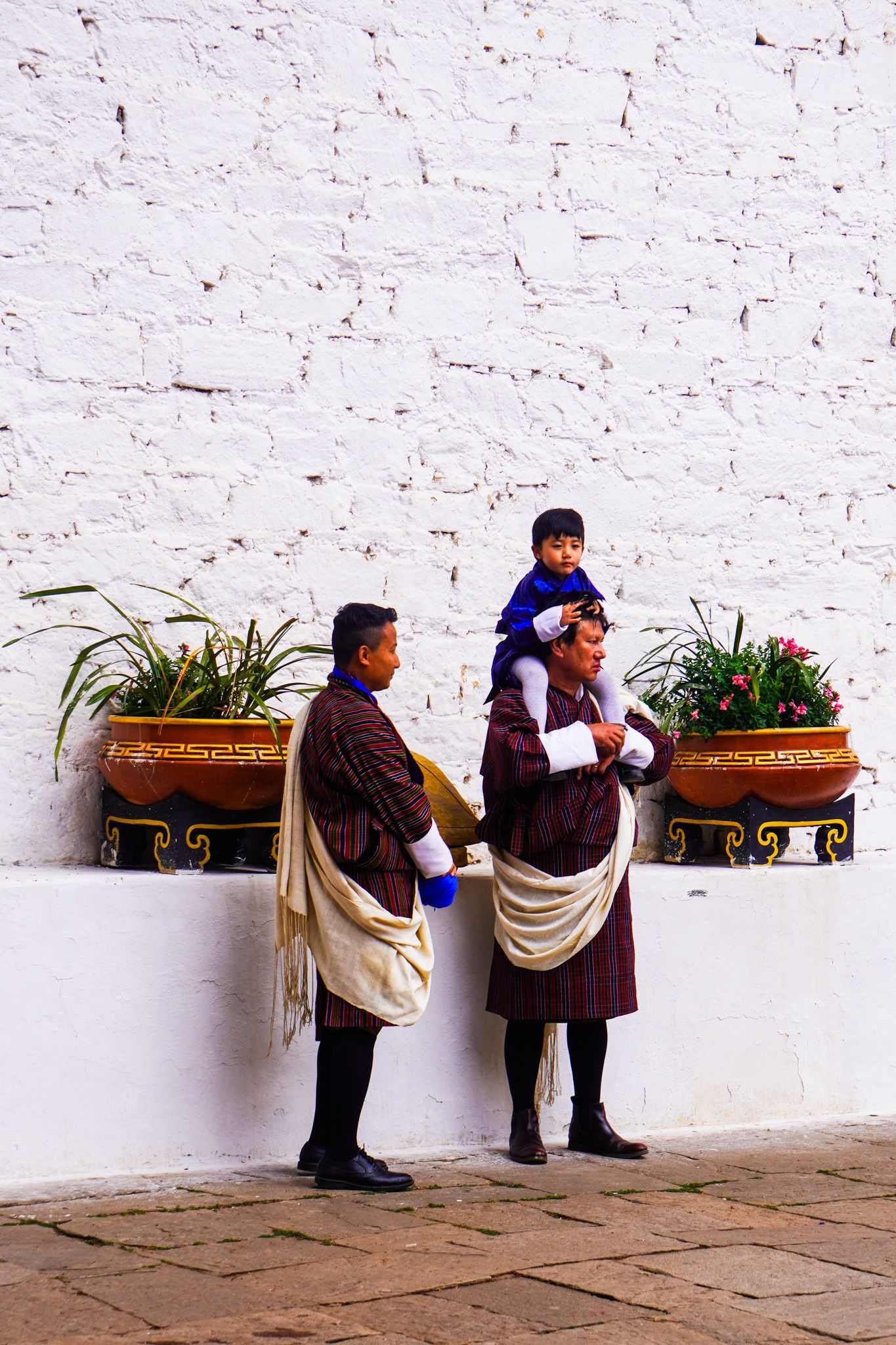 Two Bhutanese men in traditional plaid gho robes with child perched on shoulders outdoors.