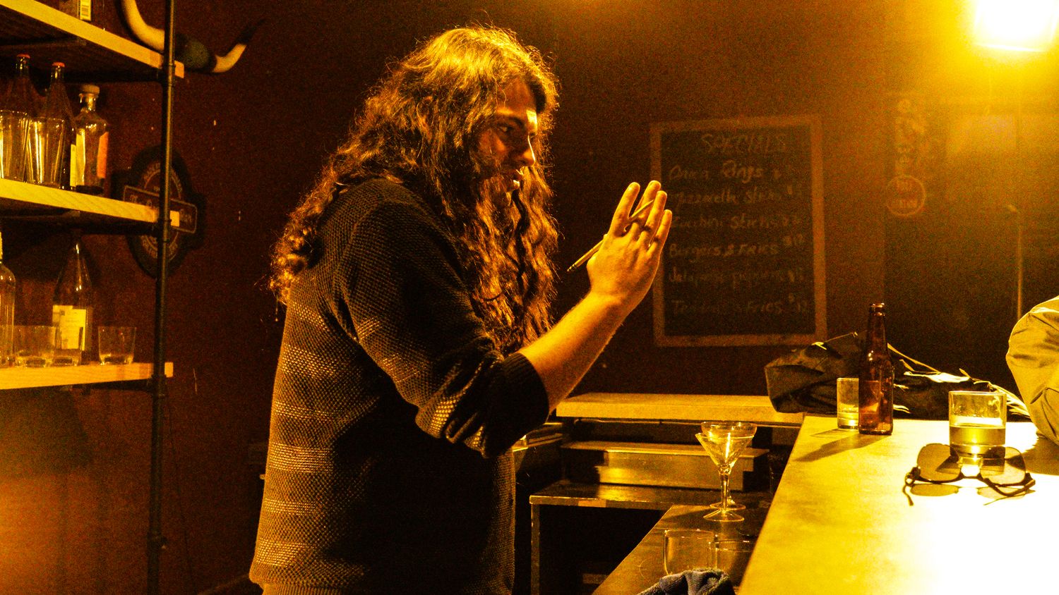 Long-haired person standing at a bar counter in dramatic warm lighting with bottles in background.