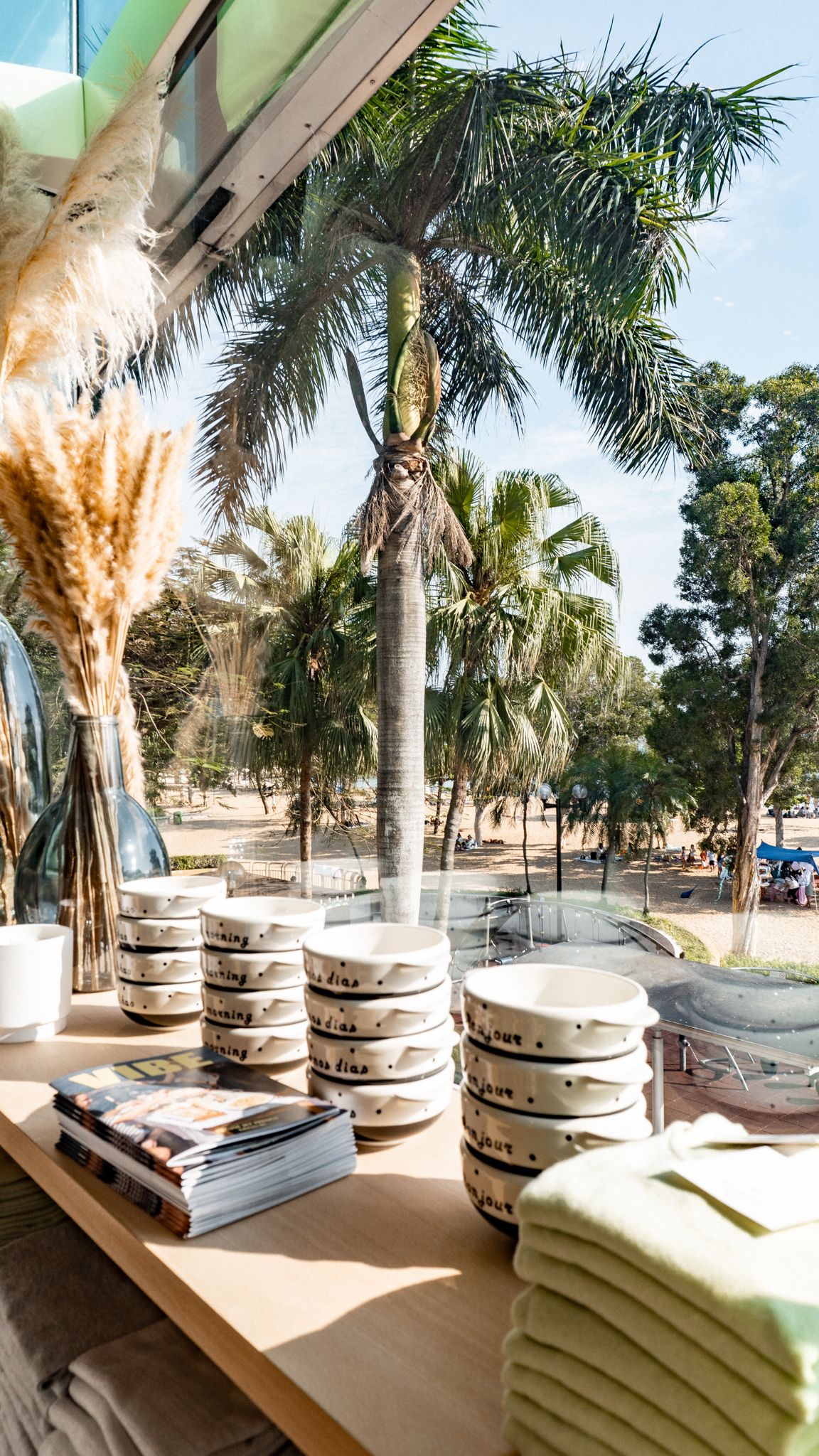 Stacked ceramic bowls and folded linens displayed on wooden shelves beside a large window overlooking tropical palm trees.
