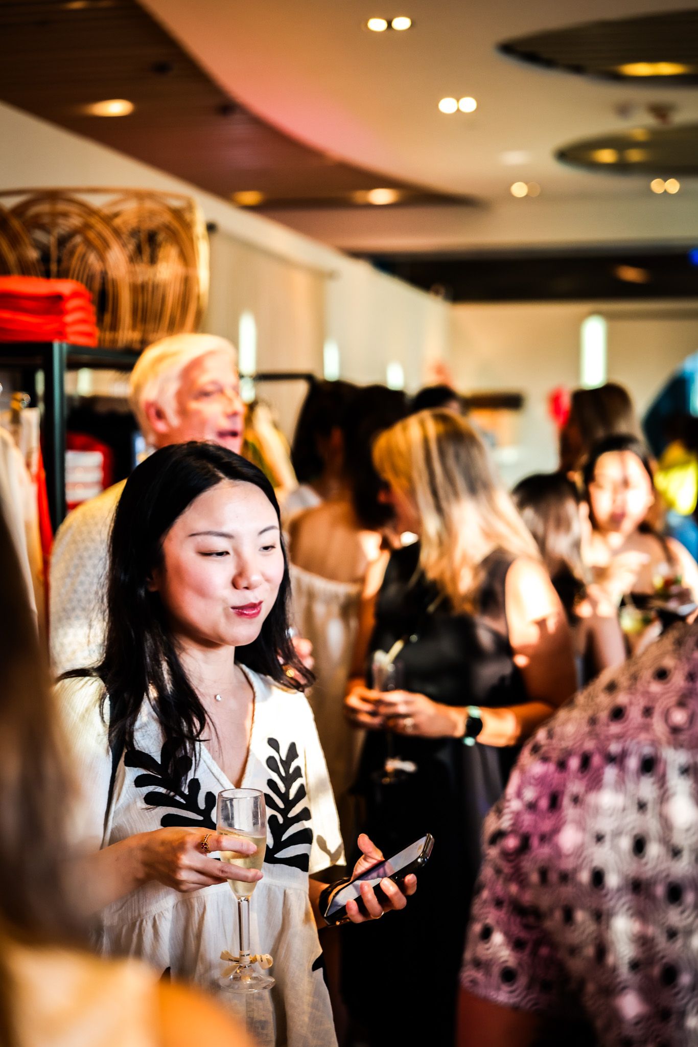 An Asian woman holds a champagne glass and phone at a busy fashion retail event or store opening.