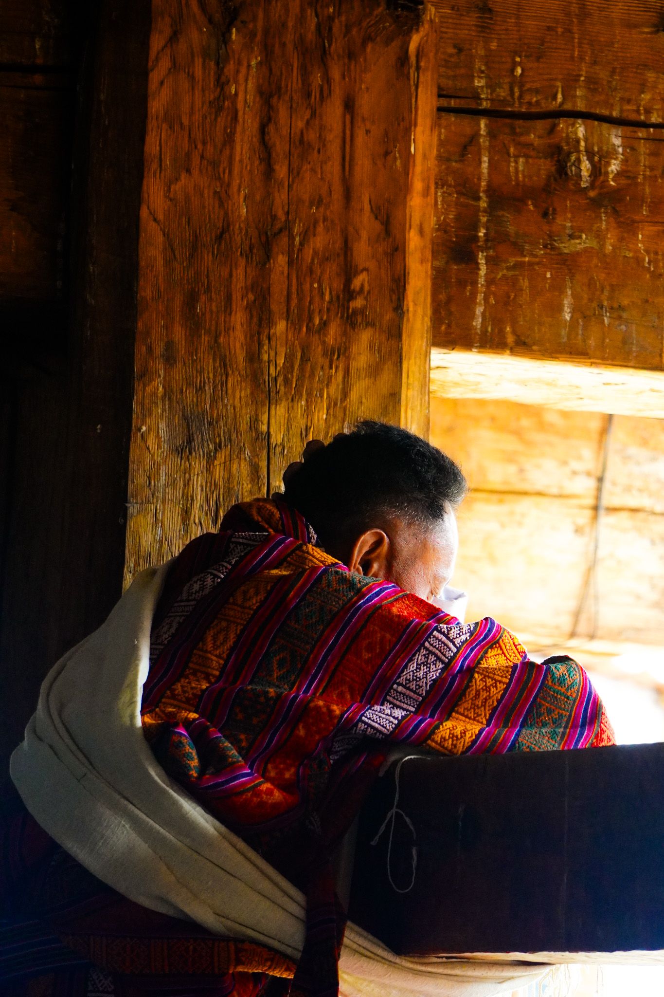 Elderly Bhutanese woman in colorful traditional dress sits by wooden window in warm light.