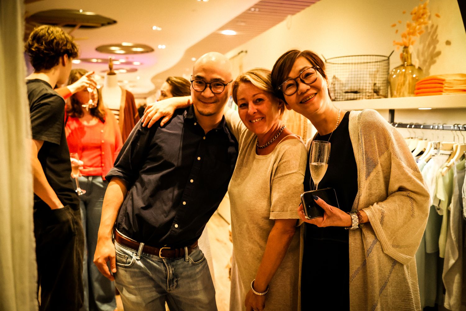 Three friends smile and pose together at a boutique store event, one holding a champagne glass.