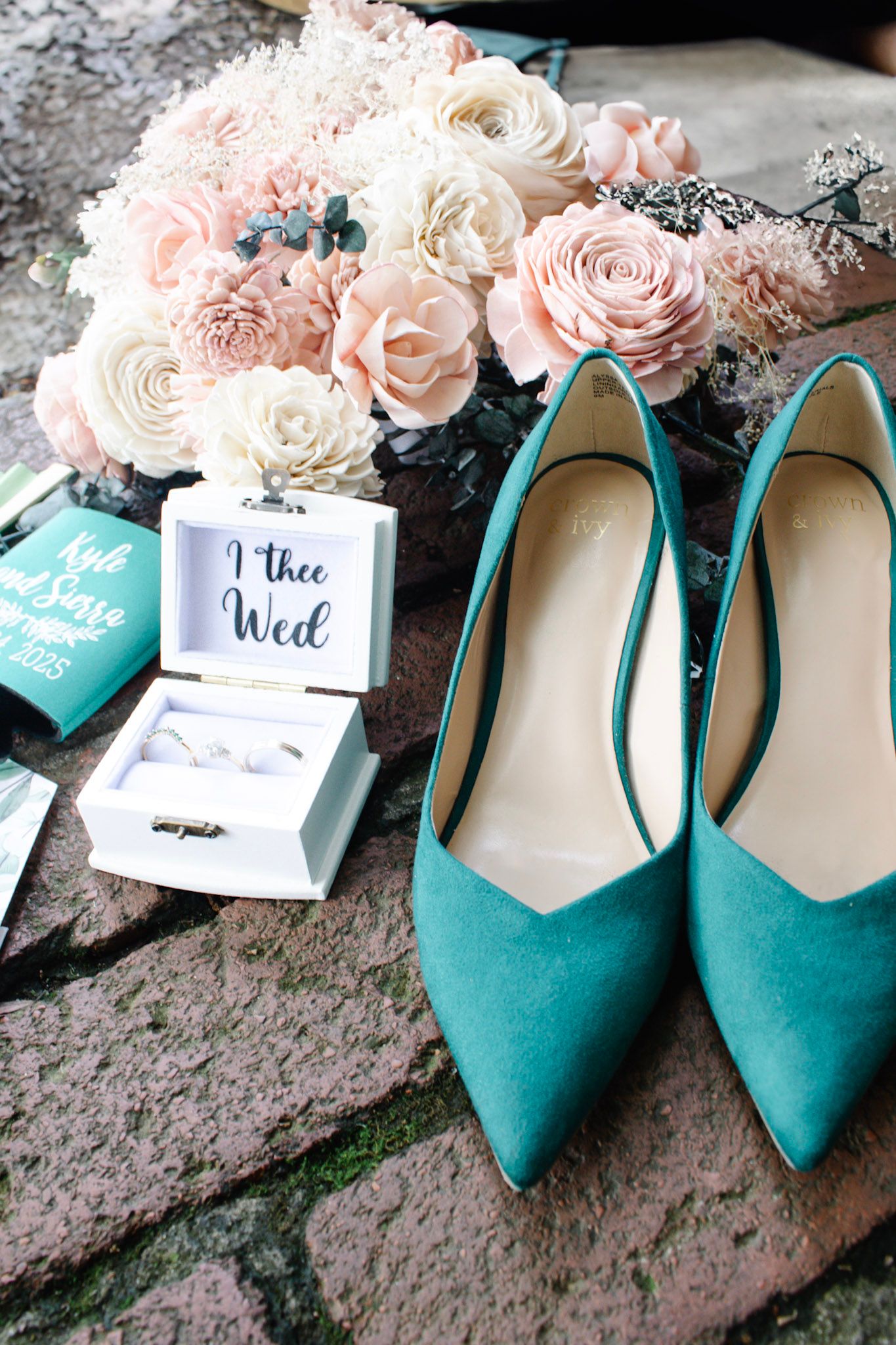 Teal suede heels beside a blush floral bouquet and I Thee Wed ring box on stone steps.