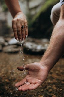 Two hands cupping and pouring fresh stream water outdoors near rocky creek bed in nature.