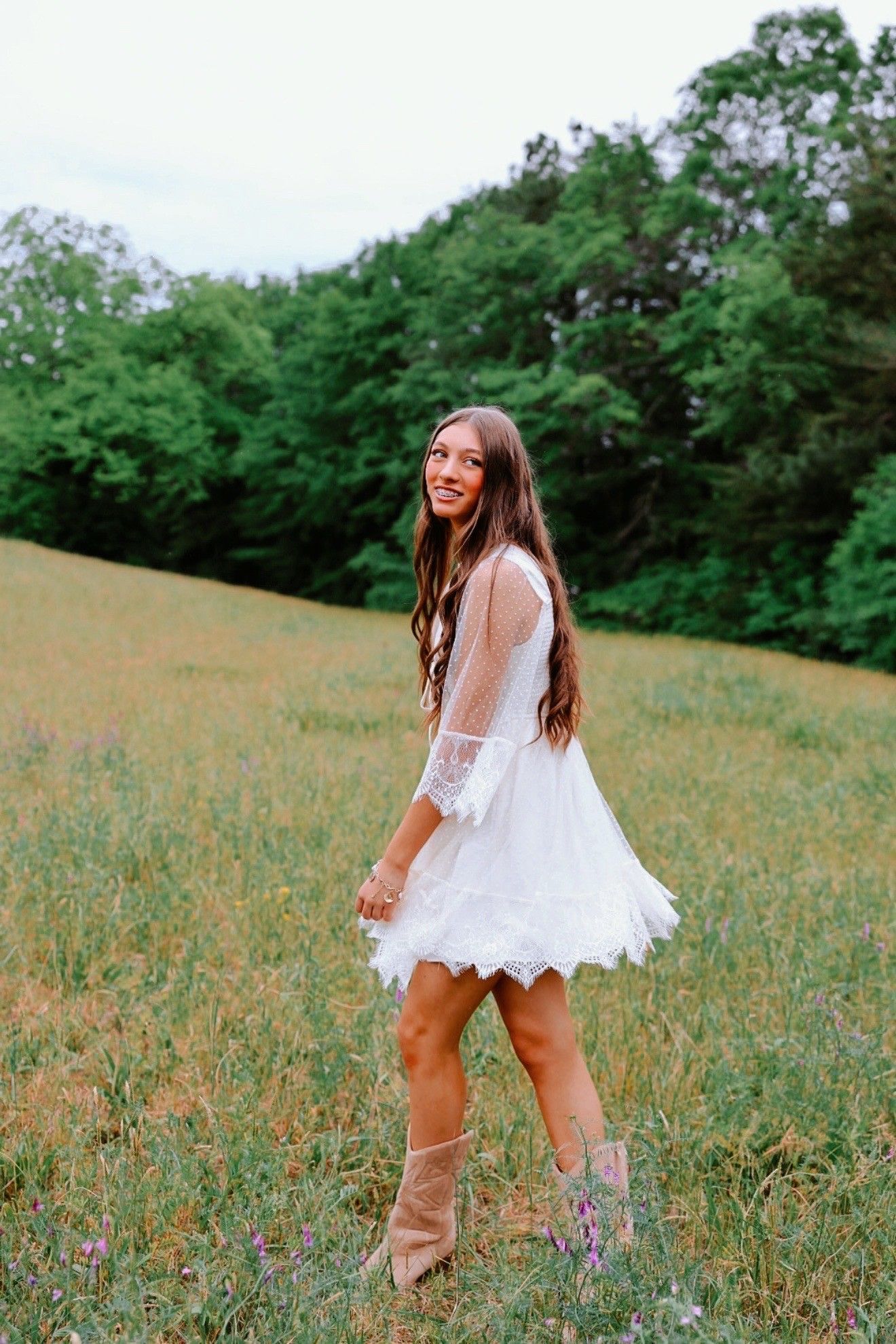 Young woman in white lace mini dress and tan boots twirling in a green meadow with trees in background.