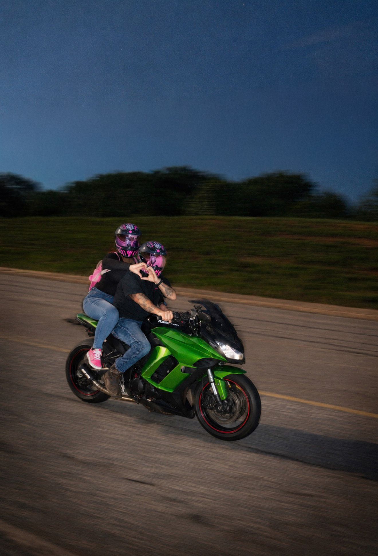 Couple riding a green Kawasaki sport motorcycle at night, both wearing pink helmets.