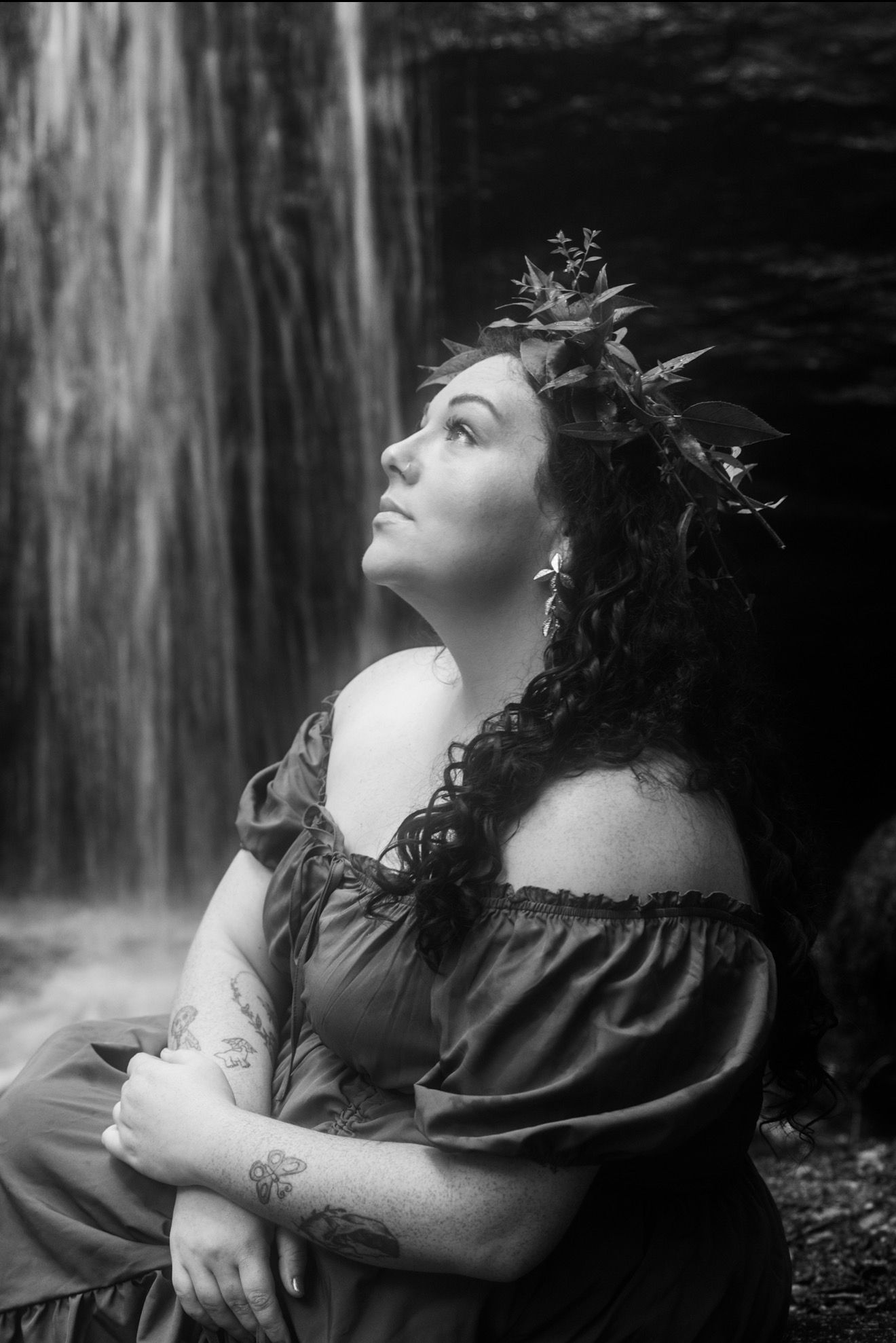 Black and white photo of a woman with curly hair and floral crown sitting before a waterfall, gazing upward dreamily.