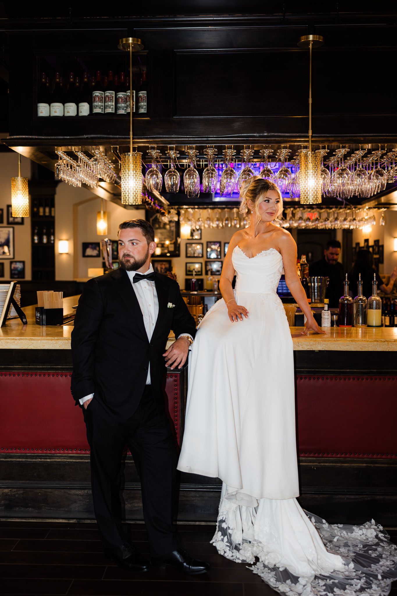 Bride in white gown and groom in black tuxedo pose elegantly at a sophisticated bar venue for their wedding portrait.