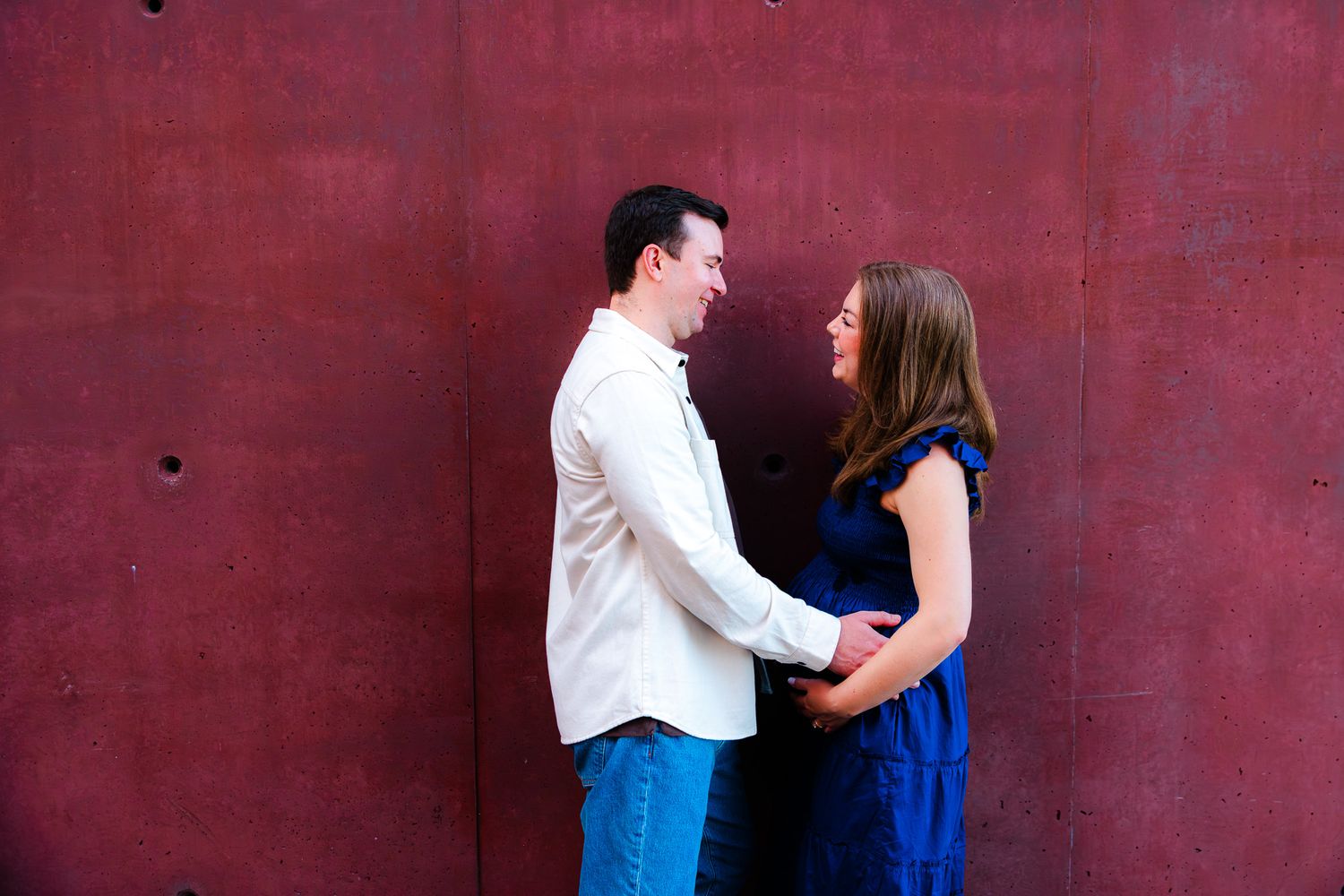 Couple holding hands and smiling at each other against a red wall backdrop.