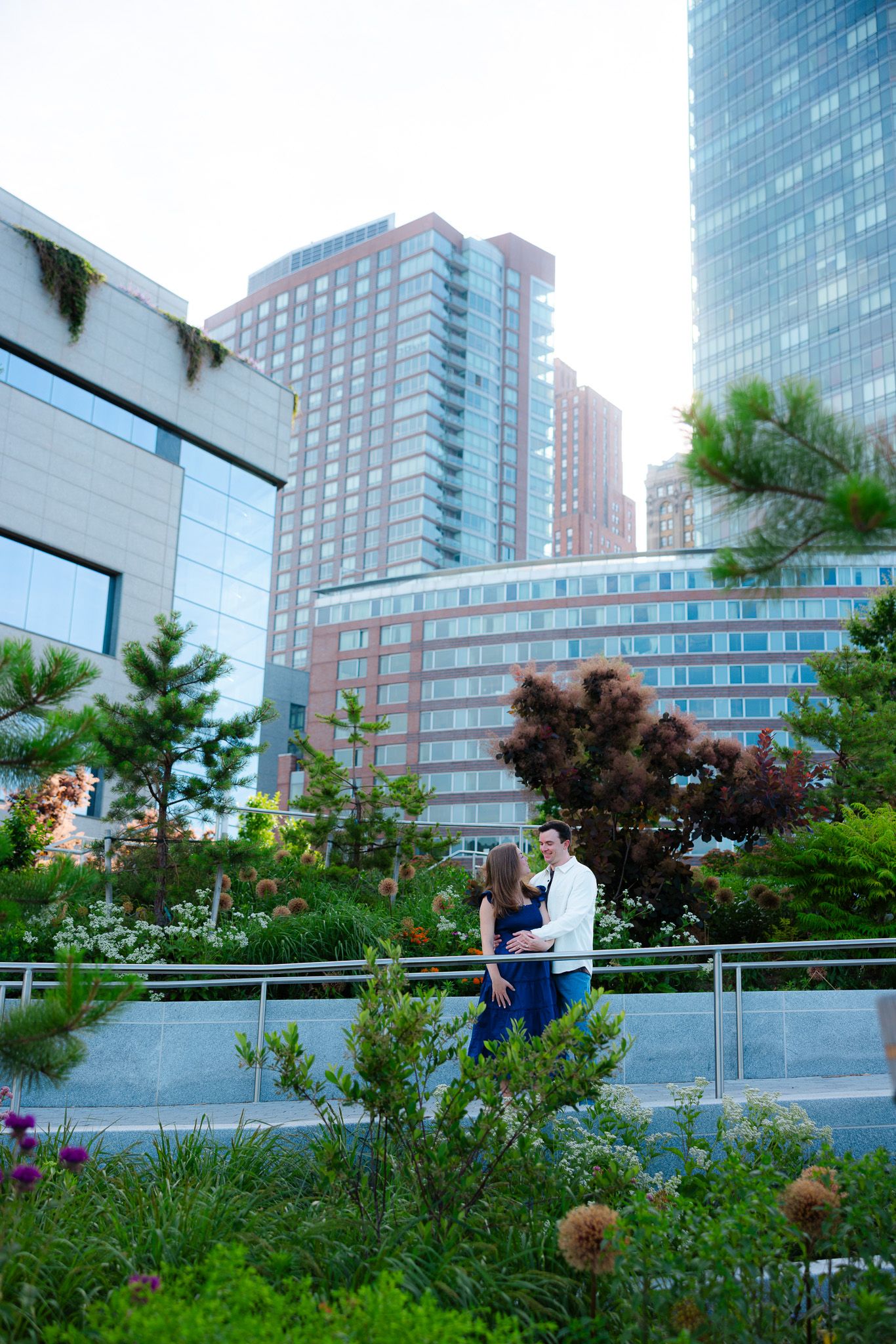 Couple embracing in an urban garden with lush greenery and modern city skyscrapers in the background.